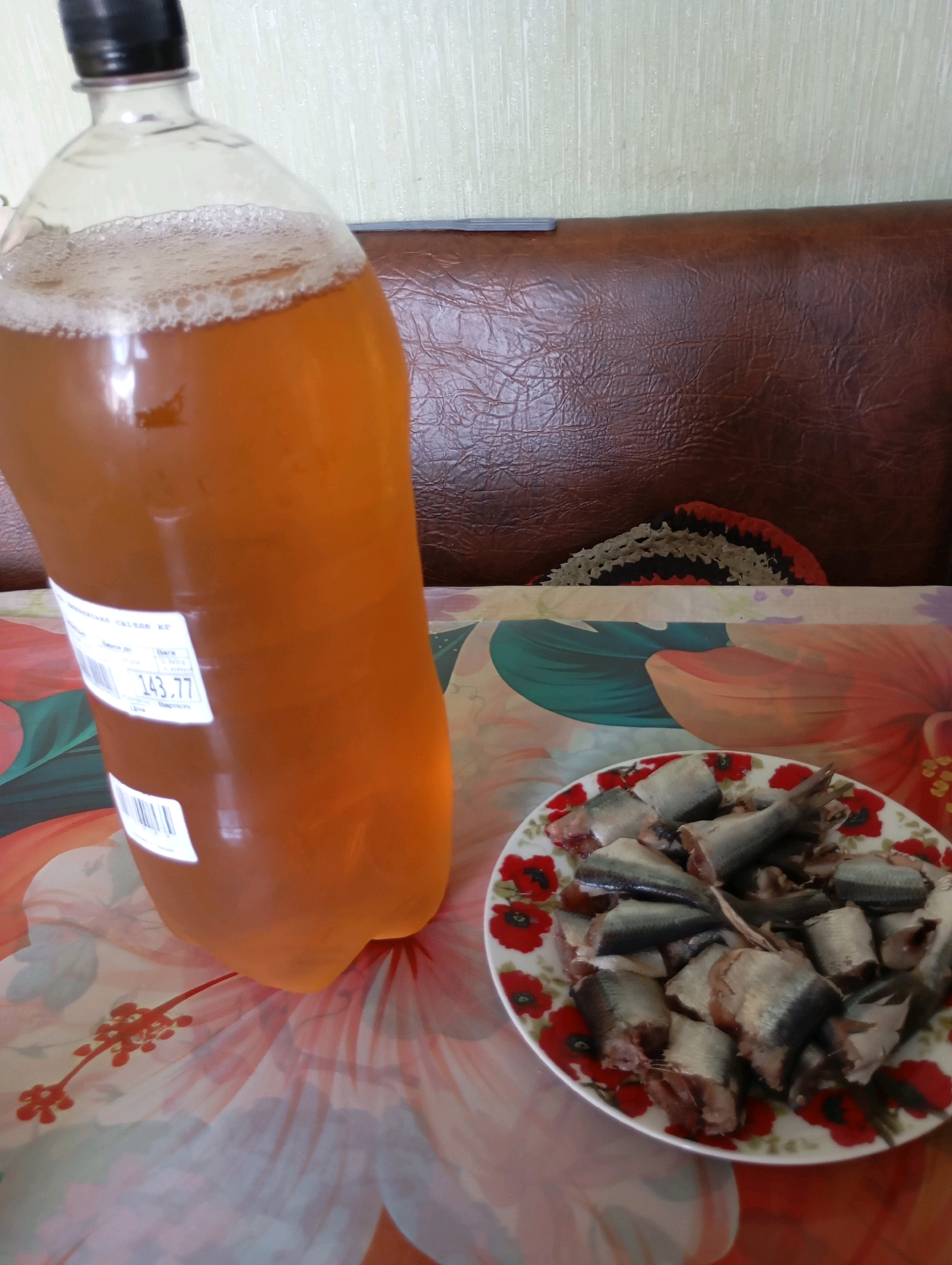 A close-up, overhead shot captures a large plastic bottle filled with amber-colored liquid, possibly beer or kvass, on a table. The bottle has a black cap and a label with Cyrillic text and numbers, including "143.77". To the right, a white plate decorated with red poppies holds numerous pieces of chopped fish, likely herring. The background features a textured brown surface, possibly a leather-like material, and a light-colored wall. A floral-patterned tablecloth is visible beneath the bottle and plate. There is no information in the image to indicate the location within the city of Horishni Plavni, Ukraine.