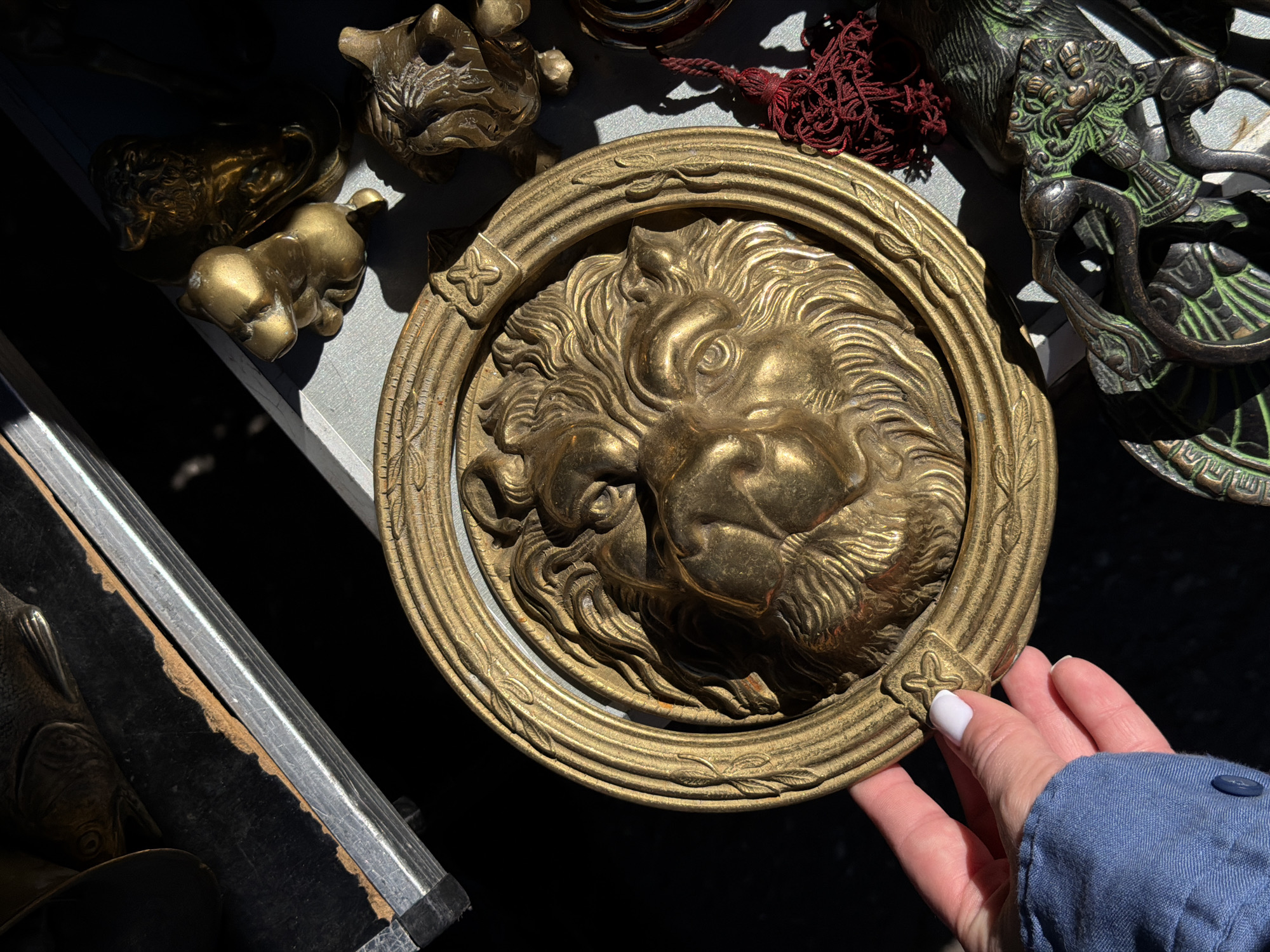 The scene depicts a close-up, top-down view of an individual's left hand holding a circular brass relief of a lion's head, likely at an outdoor market stall in Riga, Latvia. The brass relief, approximately 25-30 cm in diameter, features a highly detailed lion's head with a flowing mane, centrally placed within an ornate frame. The frame is embossed with a continuous leaf pattern and has four decorative square elements, one of which is visible under the thumb.
The hand, with fair skin and a thumb displaying white nail polish, is clad in a blue, long-sleeved garment, possibly denim or chambray, with a visible dark button on the cuff. The hand holds the lower-left quadrant of the brass relief, casting a shadow onto the object.
To the upper left, a dark, possibly wooden or laminate surface with a worn, irregular brown edge abuts a silver-colored metallic railing or frame element. In the upper right background, several smaller brass figurines are visible under direct sunlight, including a stylized, squat animal (possibly a hippo or bear cub) and a bearded bust. A dark red, multi-strand tassel rests on the surface to the lower right of the main brass relief. Below this, a section of a dark metal object with intricate green enamel or paint detailing, featuring organic or zoomorphic designs, is partially visible. The strong, direct sunlight creates high contrast and sharp shadows across the entire scene, indicating an outdoor setting.