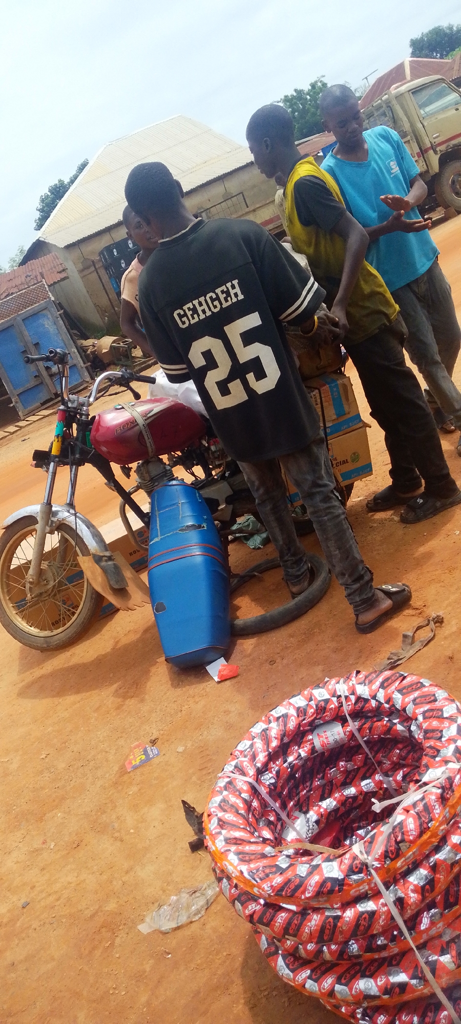 In Baissa, Nigeria, a group of young men are gathered around a red and silver motorcycle parked on a dirt road. The motorcycle has a blue plastic fuel tank attachment. One man in a black t-shirt with "GEHGEH 25" printed on the back stands facing away from the camera, while two others are partially visible. A stack of tires, wrapped in red and silver packaging with "CST" branding, is on the right side of the image. In the background, a beige truck is parked, and various buildings with corrugated metal roofs are visible. The sky is overcast.