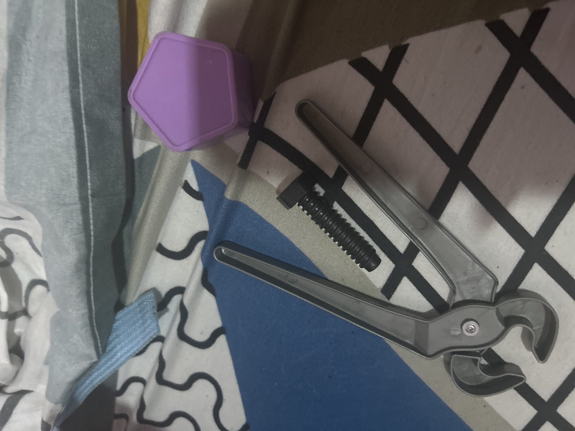 A still life photograph shows a purple pentagonal box, a black screw, and a gray plastic pair of pliers lying on patterned bedding. The background bedding has two different patterns, one featuring white fabric with wavy black lines and the other blue with white crosses. The photograph appears to be taken indoors. The location is unknown and there is no information to confirm that it is in San Juan de Los Morros, Venezuela. There are no people depicted.