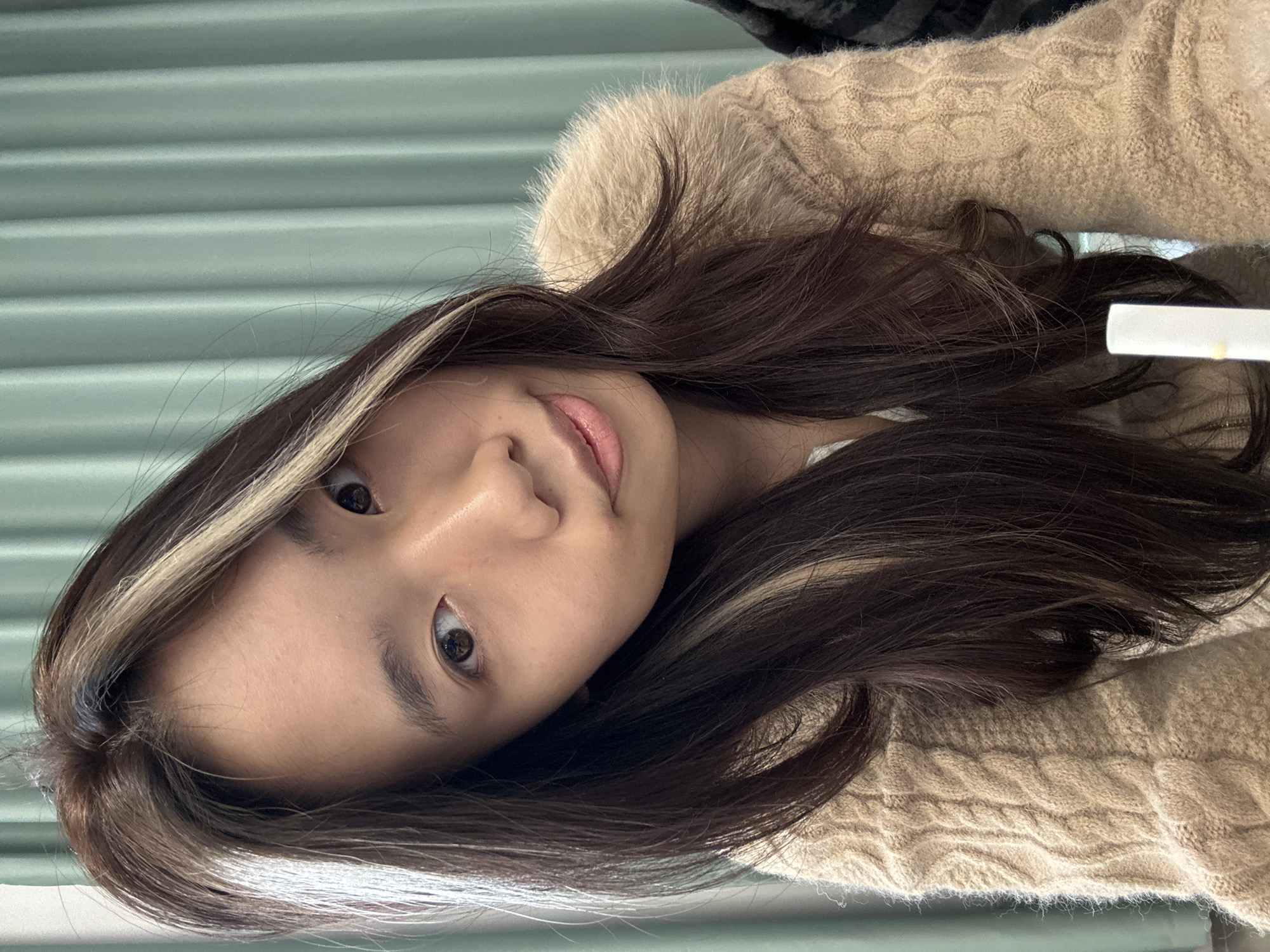 A close-up portrait depicts a young adult East Asian female, facing and looking directly at the camera, with her head tilted slightly to the viewer's left. She has long, dark brown hair with distinct light blonde streaks on the front sections. Her eyes are dark brown, and her lips are closed with a subtle upward curve. She is wearing a light brown, cable-knit sweater featuring a fluffy, possibly faux fur, collar or trim around the neck and shoulder area. A white undershirt is visible beneath the sweater at her neckline. The background consists of a uniformly pleated, light teal or pale green curtain, indicating an indoor environment. A white cylindrical object, possibly a drinking straw or a similar item, is partially visible in the lower right foreground, containing a small yellow-orange speck. The specific location in Nan'an District, China, is stated in the prompt but not visually discernible from the image itself. No other individuals or significant interactions are evident.