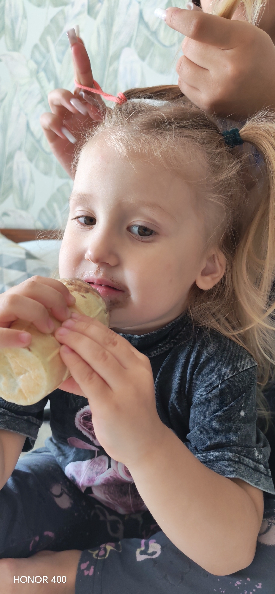 The scene is located in Noviy, Russia, depicting an indoor setting, likely a domestic residence. A young blonde-haired child, identified as a girl with brown eyes, is positioned centrally, actively eating a dark brown substance from a light-colored cone, consistent with ice cream. The dark substance is visibly smeared around her mouth and chin. Her small hands grasp the cone. She wears a dark gray, ribbed, long-sleeved shirt, possibly faded or acid-washed, with a faint pinkish-purple floral graphic on the chest. Below the shirt, patterned dark fabric, likely pants or leggings with light pink and yellow motifs, is visible.
Above the child, two adult hands with long, white-polished fingernails are present. The left adult hand holds a pink elastic hair tie, stretched around a finger and a section of the child's blonde hair, indicating the process of styling the child's hair into a ponytail. A dark teal/green elastic hair tie is already in place, securing a small ponytail on the child's head on the right side of the frame.
The background consists of light green wallpaper adorned with a repeating pattern of white tropical leaves. A portion of a patterned fabric object, possibly a pillow or blanket with geometric shapes in gray and white, is visible on the left side behind the child's head. The bottom left corner of the image contains a "HONOR 400" watermark. The scene is evenly lit, suggesting ambient indoor lighting.