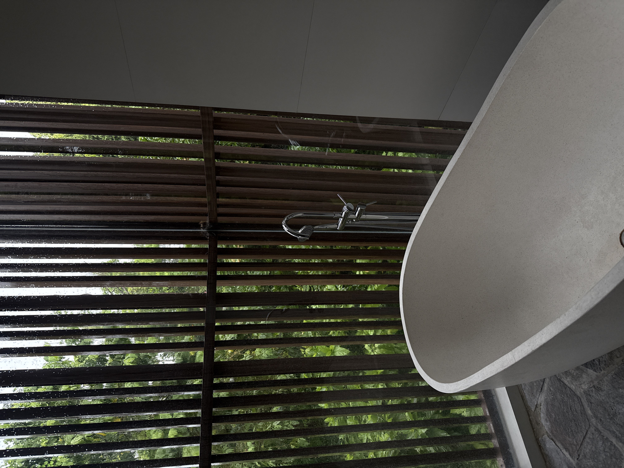 The image captures a modern bathroom interior, dominated by a light gray, oval-shaped freestanding bathtub positioned on the right side. A chrome faucet and showerhead assembly are visible near the tub. To the left, a large window or glass wall is covered by horizontal dark brown wooden slats, creating a Venetian blind-like effect. Water droplets are clearly visible on the glass and slats, indicating rain or recent rainfall. Through the gaps in the slats, lush green foliage and trees are visible, suggesting an abundant natural environment outside. The ceiling is a plain dark gray, and a textured dark stone wall is partially visible in the bottom right corner, adjacent to the bathtub. The lighting suggests an overcast or rainy daytime. The scene conveys a sense of calm and a connection with nature. There are no people or visible text.