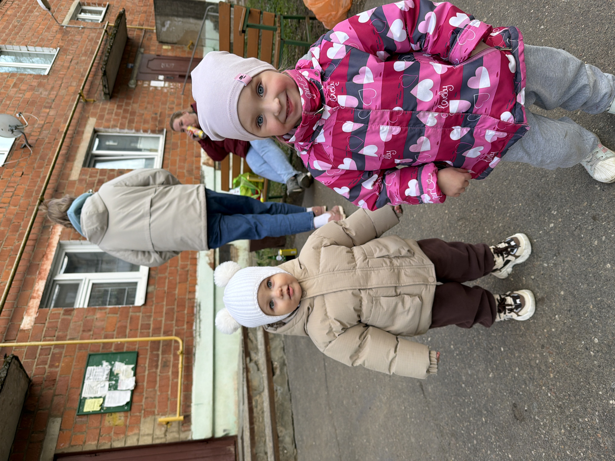 Two young children stand on pavement near a brick building in Reshetnikovo, Russia. The older child wears a pink and grey jacket with heart patterns, grey pants, and white sneakers, along with a light pink hat. The younger child wears a beige winter coat, brown pants, white sneakers, and a white hat with pom-poms. In the background, an adult in a beige jacket and blue jeans stands near a bench and another adult holding a yellow object. A notice board is affixed to the brick building. A satellite dish is visible on the building's exterior.