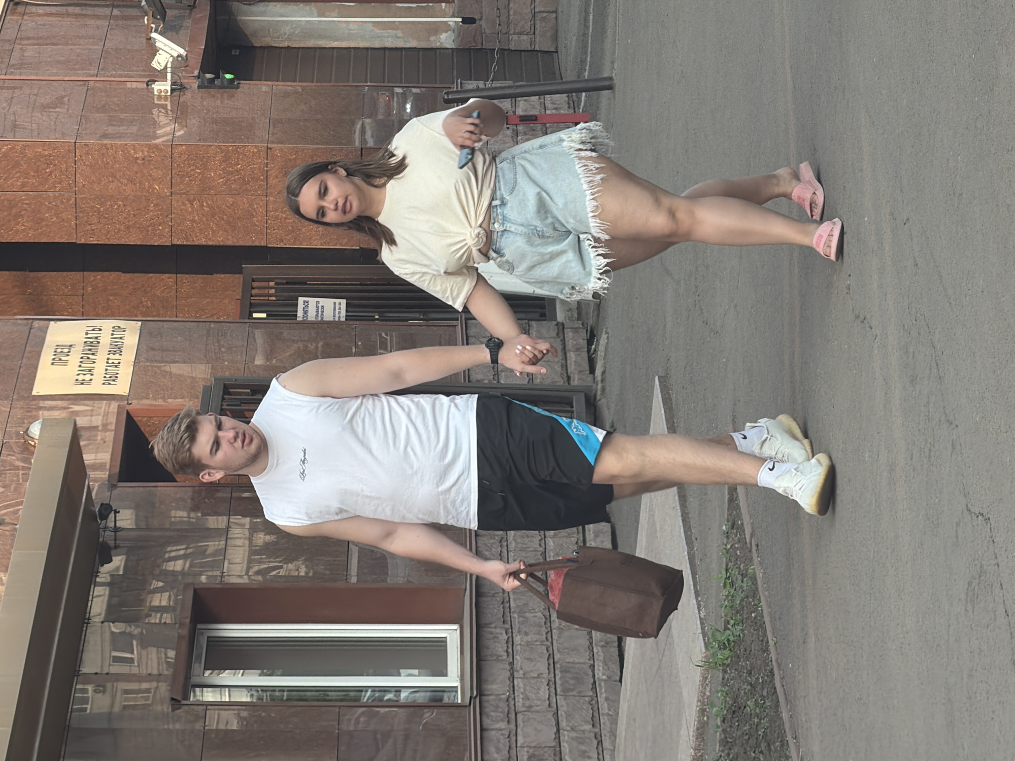 In Krasnoyarsk, Russia, a young Caucasian man and woman are walking hand-in-hand on a sidewalk in front of a building with a brown, textured facade. The man, wearing a white sleeveless shirt and black athletic shorts with a blue stripe, carries a brown tote bag in his left hand. The woman, in a cream-colored knotted t-shirt and distressed denim shorts, holds a smartphone in her right hand. They are walking away from the camera, towards the right side of the frame. The building has a modern design with large windows and signage indicating it is a public space. Traffic lights and security cameras are visible on the building's exterior. The ground is paved with asphalt.
