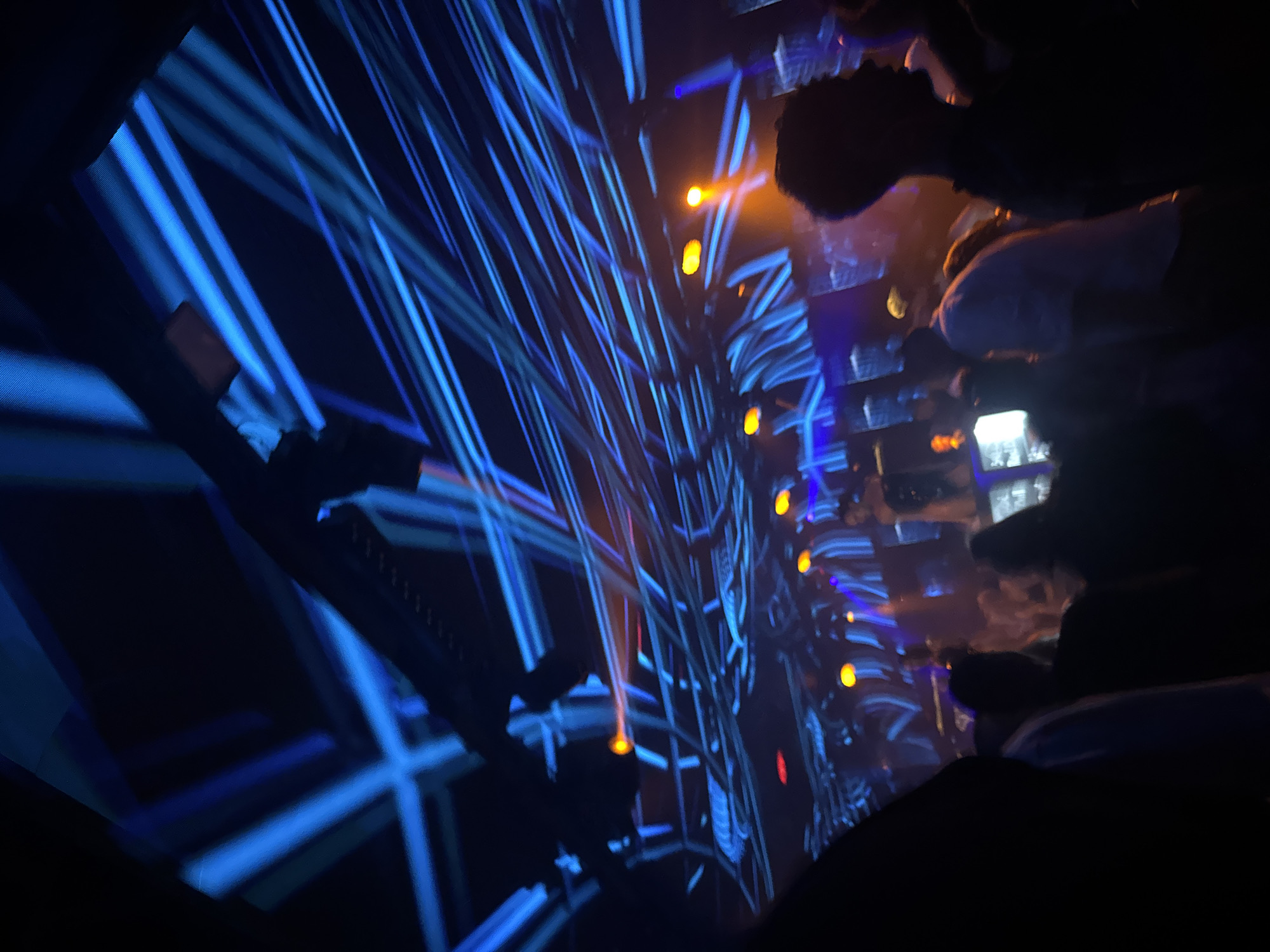 The image shows a dark indoor space with a complex lighting display. Multiple bright blue light beams are crisscrossing the scene, forming geometric patterns. Interspersed among the blue lights are several small, orange lights. Some blurred figures are visible in the foreground, suggesting the presence of people observing the light show. The specific location within the city of Unknown, Unknown cannot be determined from the image alone.