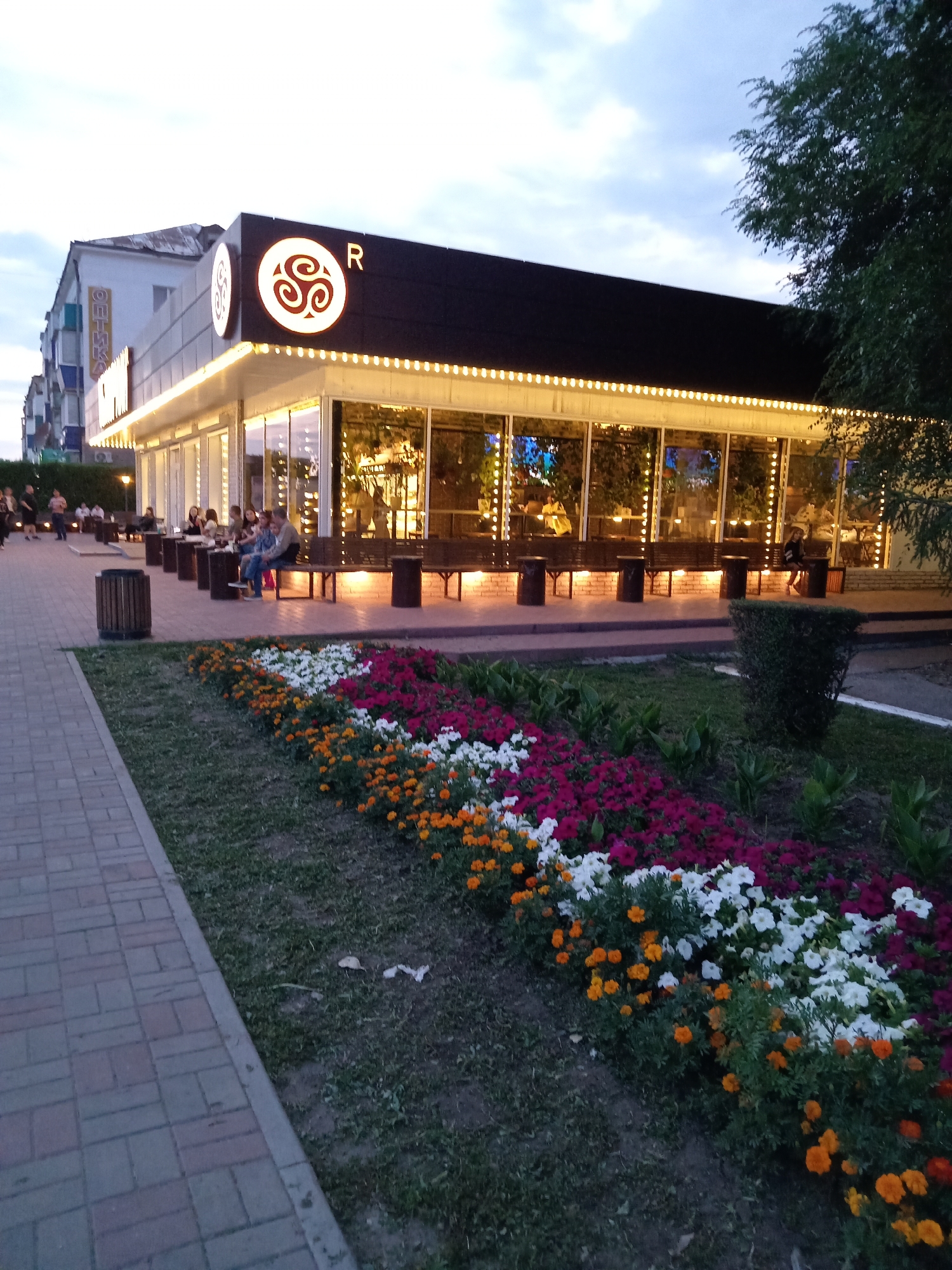 The image shows an outdoor seating area of a building, likely a cafe or restaurant, in Rudny, Kazakhstan. The building has a modern design with large glass windows and a dark facade adorned with string lights. A prominent circular logo with a swirling design and the letter "R" is visible on the front of the building.
In front of the building, several people are seated at tables and on benches, appearing to be patrons enjoying the evening. A paved walkway leads to the seating area, and a well-maintained flower bed with colorful flowers in shades of orange, white, and pink borders the walkway and the building. A cylindrical trash bin is also visible near the walkway.
The surrounding environment suggests an urban setting with other buildings visible in the background and trees providing some shade. The sky is overcast, indicating it's either early morning or late evening. The overall impression is of a pleasant and inviting outdoor public space.