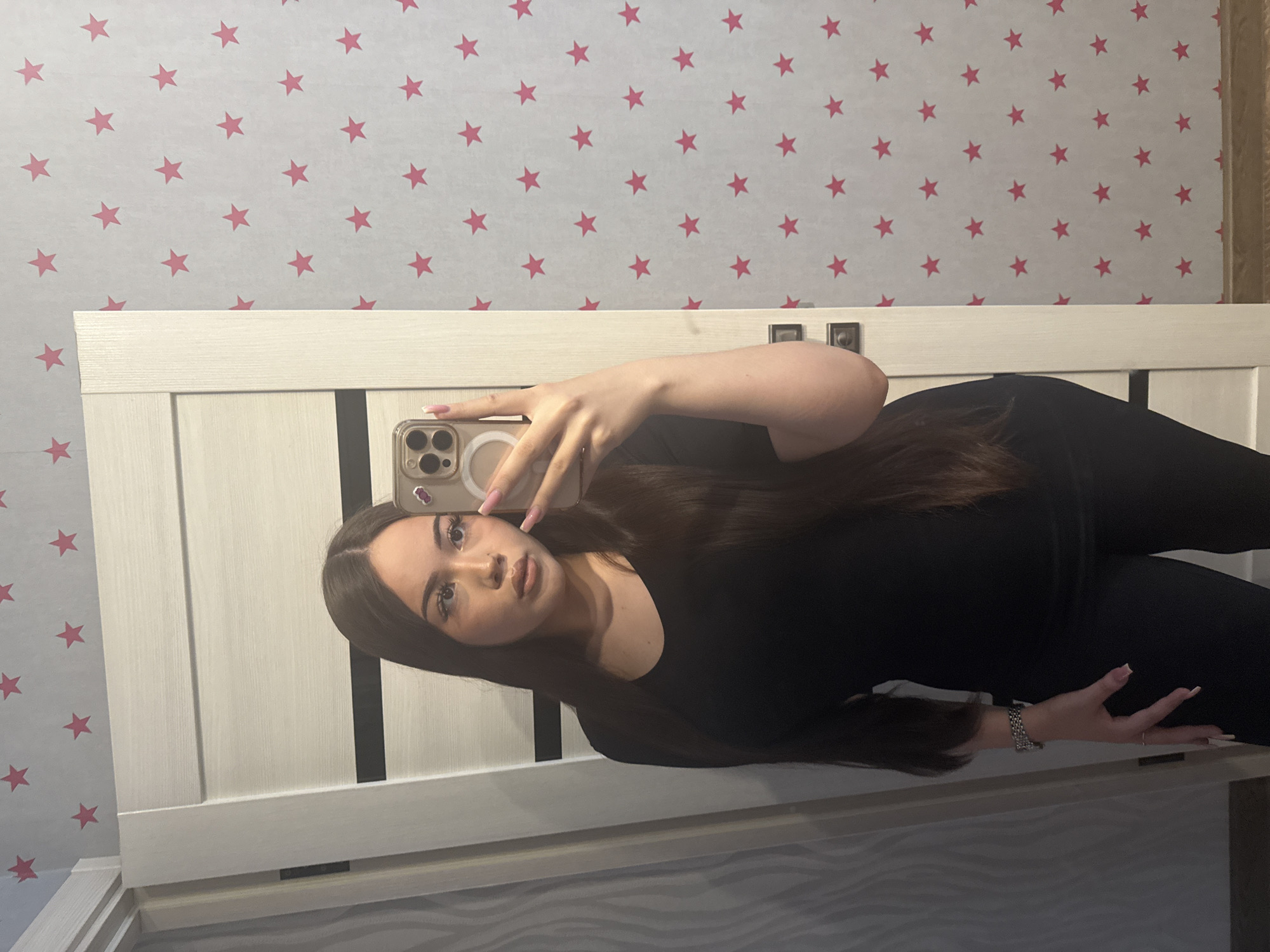 A young woman is captured in a mirror selfie, positioned diagonally across the frame, looking directly into the camera's reflection. She has long, straight dark hair, parted in the middle, and is wearing a plain black top. Her face features defined eyebrows, eyeliner, and what appears to be lip gloss. Her fingernails are manicured with a light pink polish.
She holds a smartphone, likely an iPhone with a triple-lens camera array and a white MagSafe ring visible on its case, in her right hand, extended towards the mirror. On her left wrist, she wears a textured, silver-colored bracelet, and a simple ring is visible on a finger of her right hand.
The background features a light-colored wooden bed frame or headboard with vertical black accents. Above this, a light grey or white wallpaper adorned with numerous scattered pink stars creates a distinct pattern. Below the headboard, a floor or rug with a wavy, light grey pattern is visible in the reflection. The scene is brightly and evenly lit, characteristic of an indoor setting. No visible text is present in the image.