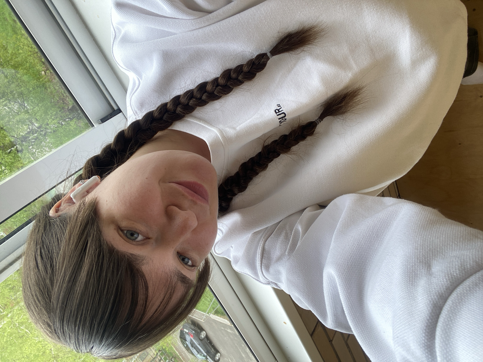 A person is positioned by a window, likely indoors. They are wearing a white sweatshirt and have brown hair styled in two braids. An earbud is visible in their left ear. Through the window, a glimpse of an exterior scene includes a building and a parked car. It is not possible to definitively confirm the location as Votkinsk, Russia, from this image alone.