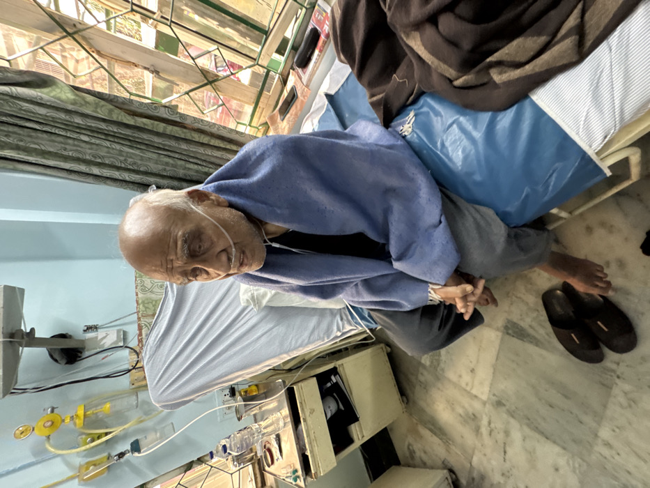 An elderly man rests in a hospital bed, wearing a blue blanket and an oxygen tube in his nose. His bare feet are near a pair of dark slippers. The bed is equipped with medical equipment and tubes. A window is visible with sunlight filtering through. The floor is marble.