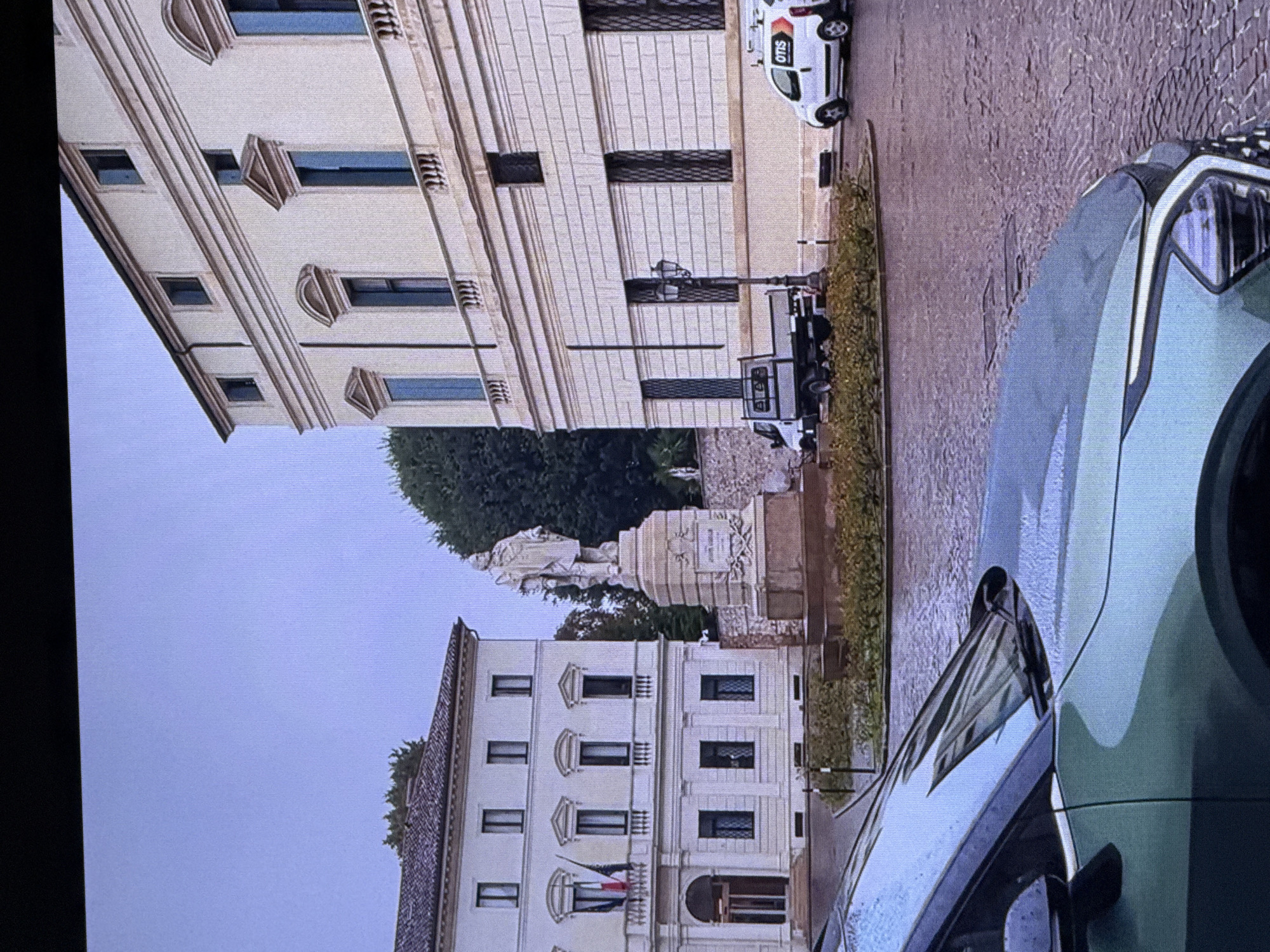 The image shows a street scene in front of a large, ornate building. There is a statue in the foreground, and a car parked on the side of the road. A small white van with "OTIS" written on it is also visible. The sky is overcast, and the ground is wet, suggesting it may have recently rained. The architectural style of the buildings and the presence of palm trees indicate a location in a warm climate, possibly the Mediterranean or the Middle East. However, without additional context, it is not possible to definitively identify the city as Dubai, United Arab Emirates.