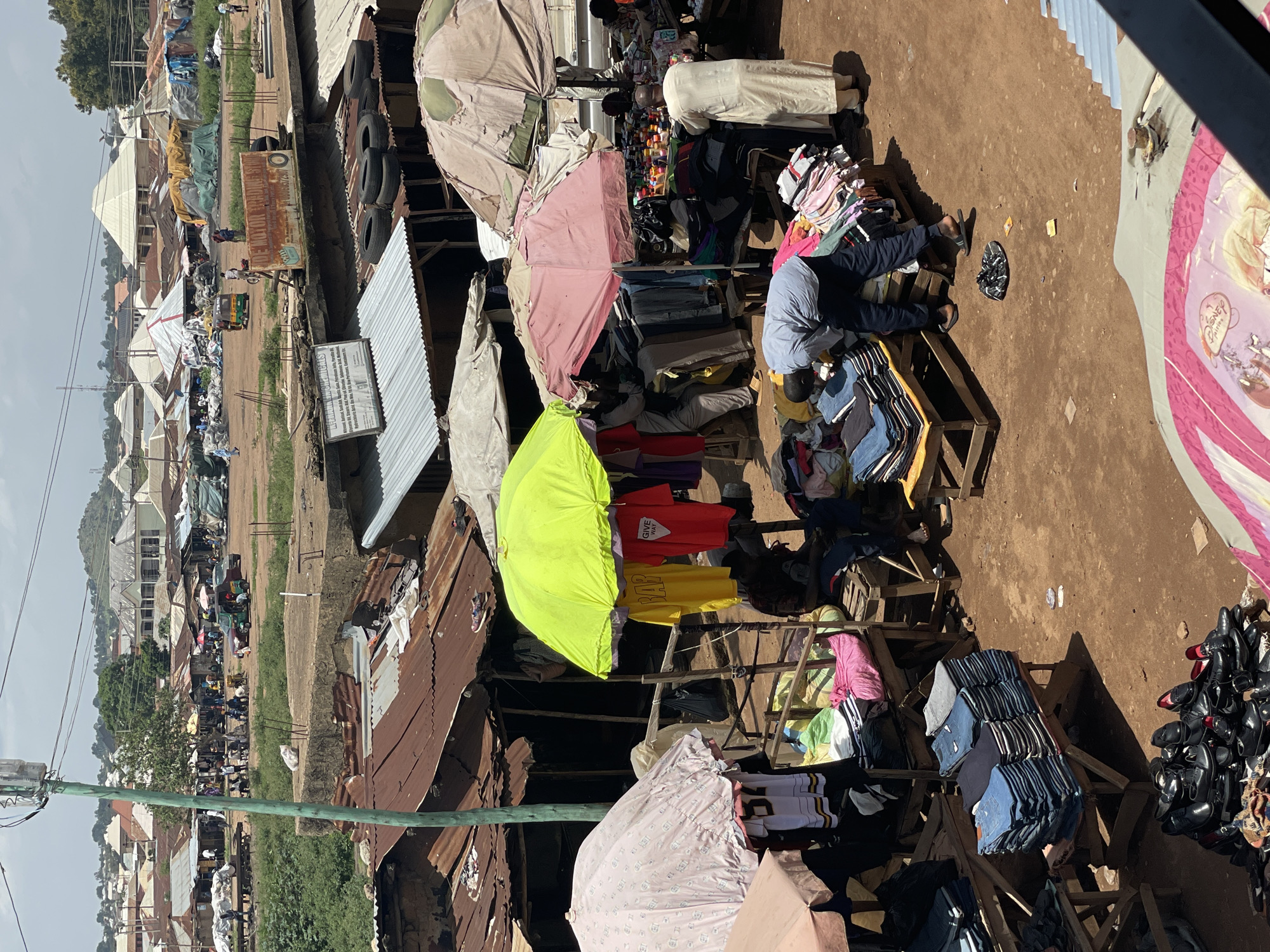 This is an eye-level, medium shot of an outdoor market scene. In the foreground, a variety of clothing is displayed on wooden tables and stands, with stacks of folded jeans and other garments. A pile of black shoes is also visible. Several people are browsing or working at the stalls. A man in a grey shirt and dark pants is bent over a display of clothes, and another man in a long, light-colored robe stands to his right, facing away from the camera. Two large, colorful umbrellas, one bright green and one pink, provide shade over some of the stalls. In the background, more market stalls with corrugated metal roofs are visible, along with a few vehicles, including a tricycle. Beyond the market, buildings and a distant, hilly landscape can be seen under a cloudy sky. The ground is dirt.