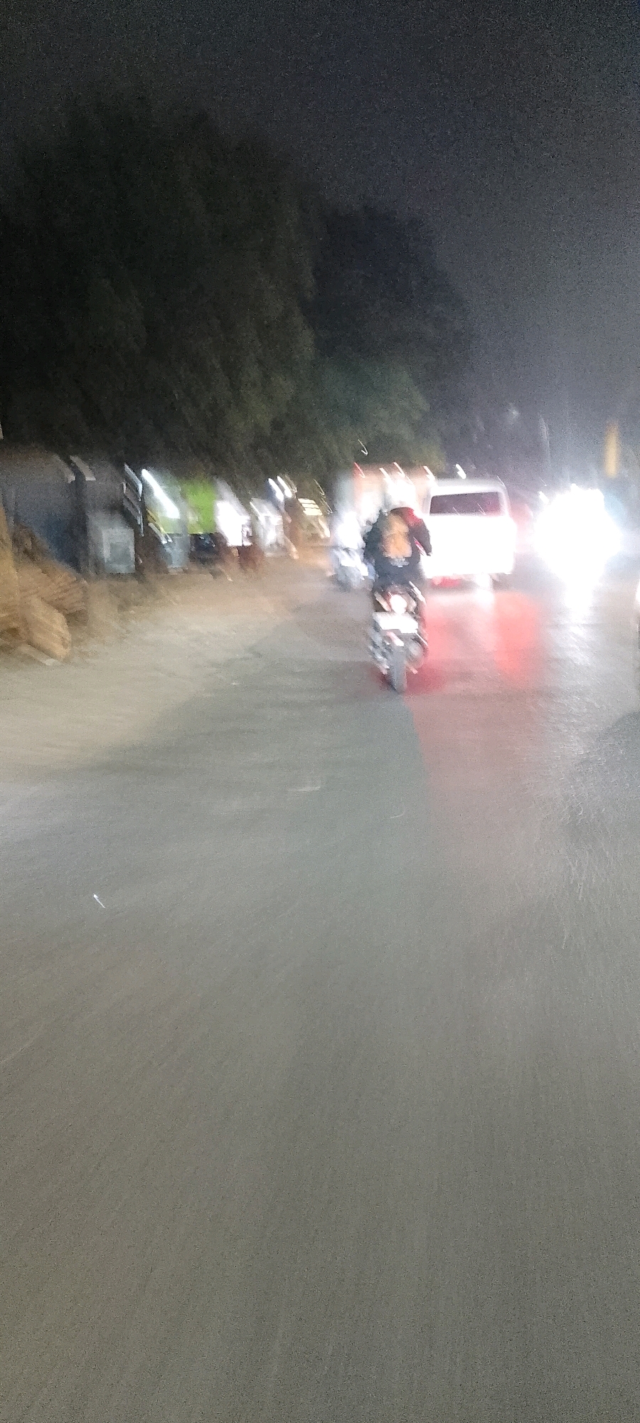 A blurry, nighttime image shows a street scene in Bekasi, Indonesia. A person rides a motorcycle in the foreground, facing away from the camera. The motorcycle's headlight and taillight are visible. In the background, several vehicles, including a white van, are illuminated by their headlights. Trees line the left side of the road. The road appears wet, possibly from rain.
