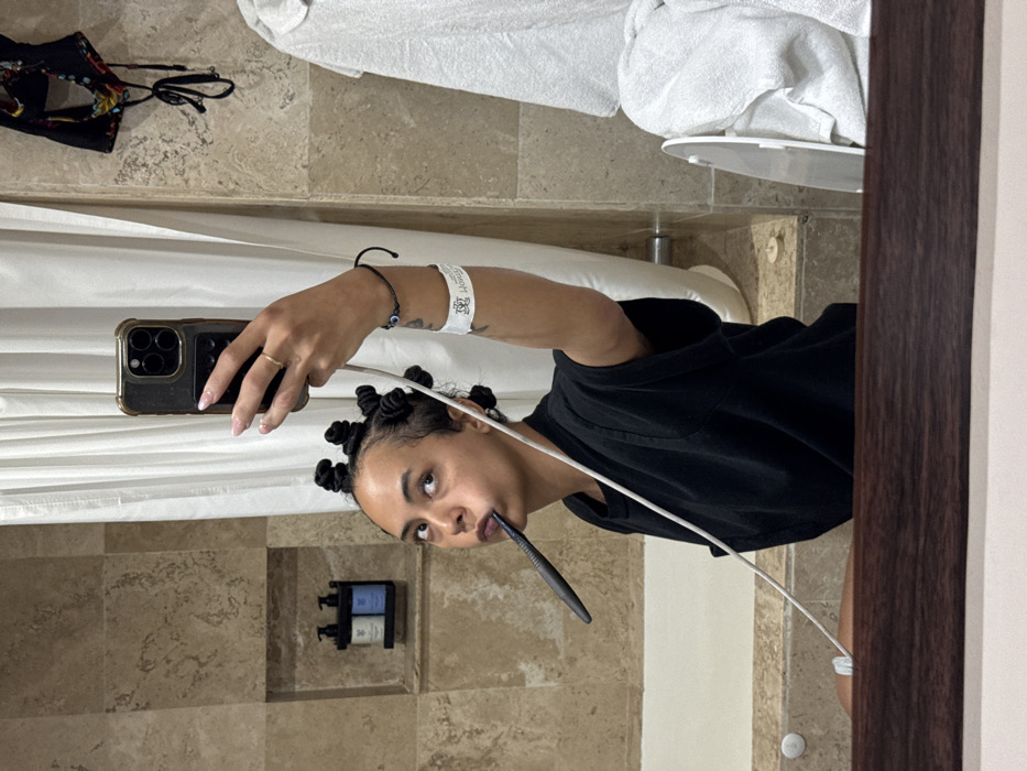 This image depicts a selfie taken in a bathroom mirror at Hotel Montetaxco in Taxco, Mexico.
The subject is a person with dark skin and dark hair styled into multiple small coiled buns. They are wearing a black short-sleeved top. A black toothbrush is horizontally positioned in their mouth. The person holds a dark-colored smartphone, likely an iPhone with three camera lenses, in their right hand to capture the mirror reflection. Their right hand displays light pink nail polish and a gold-colored ring on the ring finger. A white charging or audio cable extends from the phone, draping across their body. On their right wrist, a white wristband with the "MONTE TAXCO" logo is visible, along with a black string bracelet featuring a blue evil eye bead. The subject's gaze is directed upwards and to the right, towards the phone's camera.
The bathroom features walls tiled with light brown, mottled natural stone. A white bathtub is partially visible in the lower left quadrant of the image. Above the bathtub, a recessed wall niche contains two dark pump bottles with light blue and light green labels. A white fabric shower curtain hangs to the right of the bathtub. To the upper right, a black fabric item with a colorful pattern, possibly swimwear, hangs on the wall. A white towel is draped over an unseen object on the lower right, adjacent to a partially visible white circular fixture. The bottom edge of the mirror is framed by a dark wooden border.