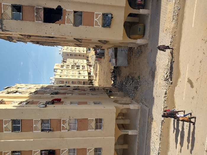 A cityscape reveals multi-story, tan-colored buildings with repetitive window designs, some damaged, hinting at conflict or neglect. A street runs alongside one building, featuring a group of people and their shadows. Debris and tents are visible between the buildings. The perspective is somewhat distorted, suggesting the photo was taken from an unusual angle or location. The sky is clear and blue.