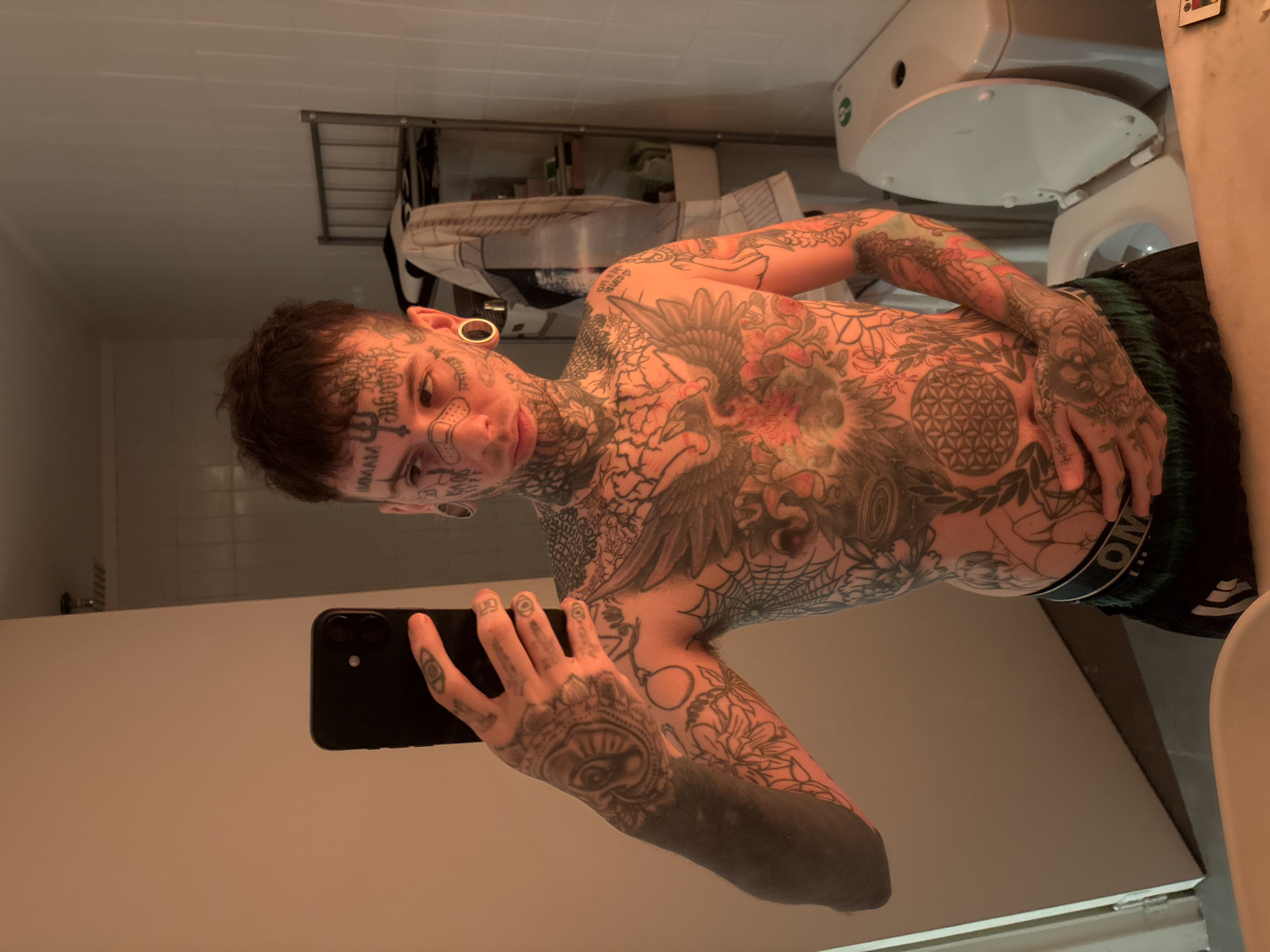 A man with extensive tattoos covering his face, torso, arms, and hands takes a selfie in a bathroom mirror. He's wearing dark shorts with a visible waistband. The bathroom includes a toilet, a rack above the toilet, and white walls with tiled accents. The location is not discernible as being in Buenos Aires, Argentina.
