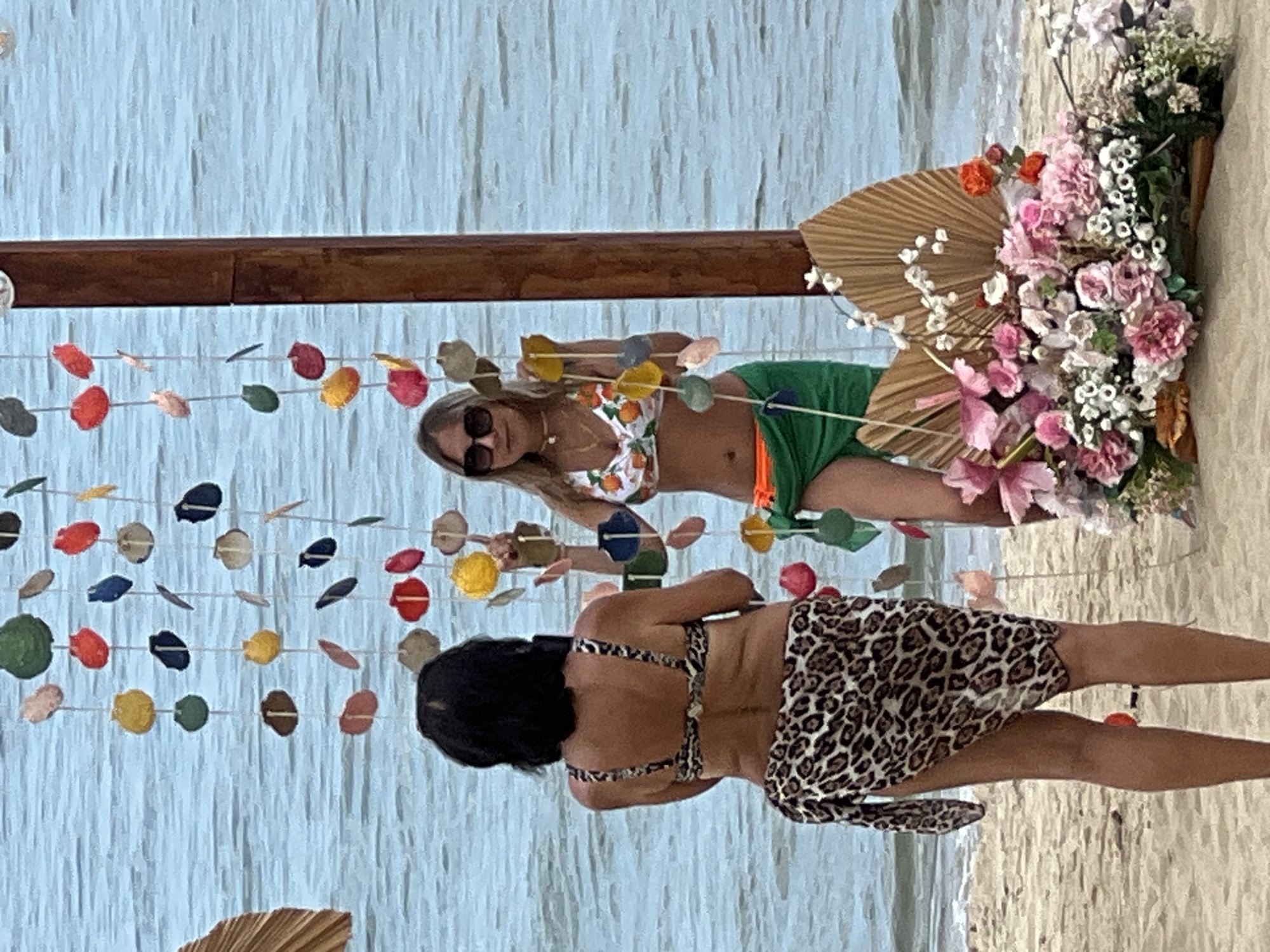 Two women are posing for a photo on a sandy beach in front of the ocean in Nha Trang, Vietnam. The woman on the left is wearing a leopard print bikini top and a matching sarong. The woman on the right is wearing an orange and white fruit-patterned bikini top and a green sarong. Both women are facing away from the camera and looking towards something off-frame to their left. Behind them, a wooden post supports a string of colorful seashells and other decorative items, creating a festive backdrop. A large arrangement of tropical flowers and dried palm leaves is placed on the sand to the right of the women, adding to the celebratory atmosphere. The ocean is calm and blue, with gentle waves lapping at the shore. The sky is overcast.