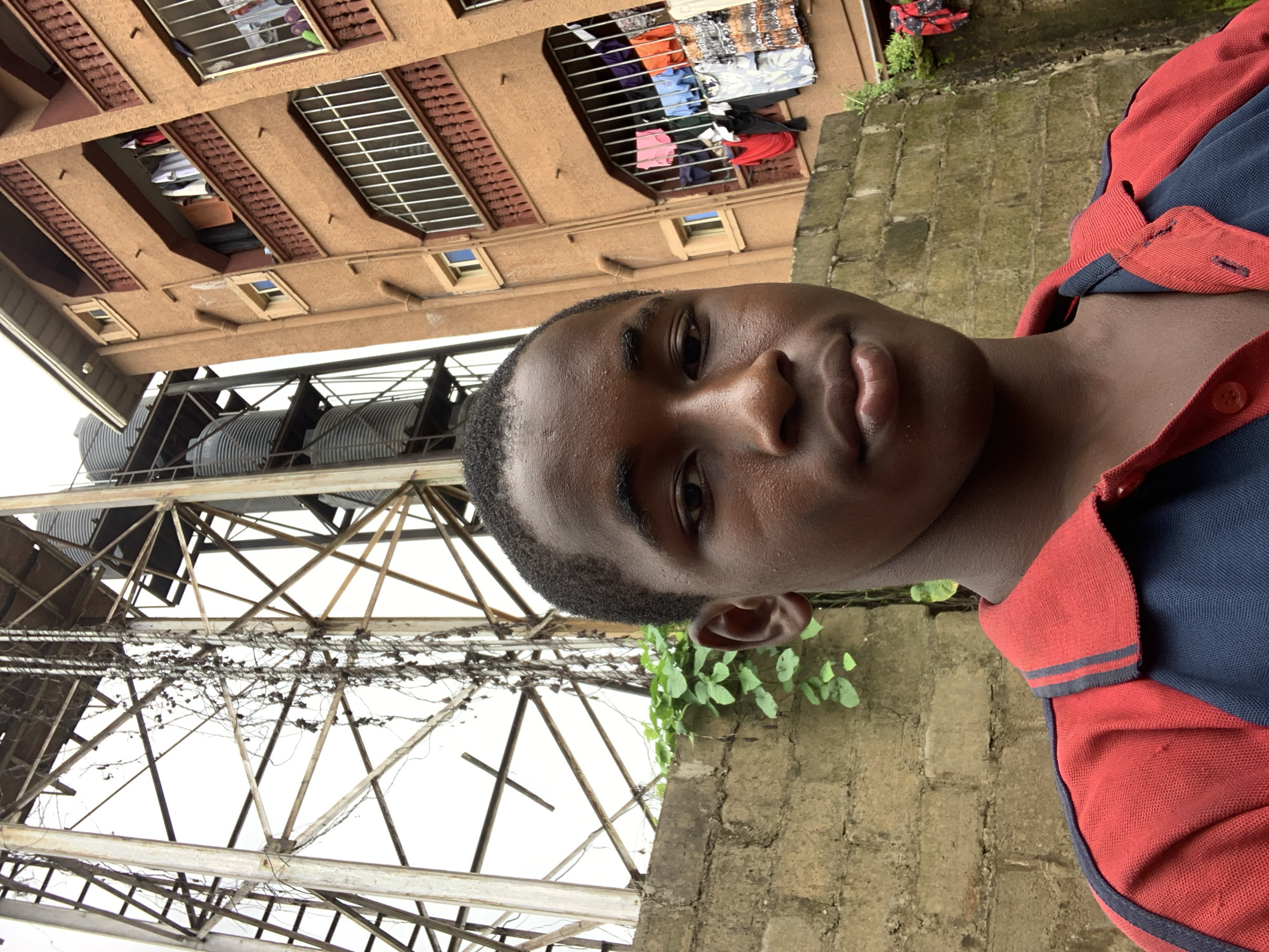 A young man with dark skin and short, dark hair takes a selfie in Nkpor, Nigeria. He is wearing a red and dark blue collared shirt. The background shows a brick wall, green foliage, and a large, industrial metal structure with water tanks, likely a water tower, with an adjacent brown building with multiple balconies where laundry is hanging.
