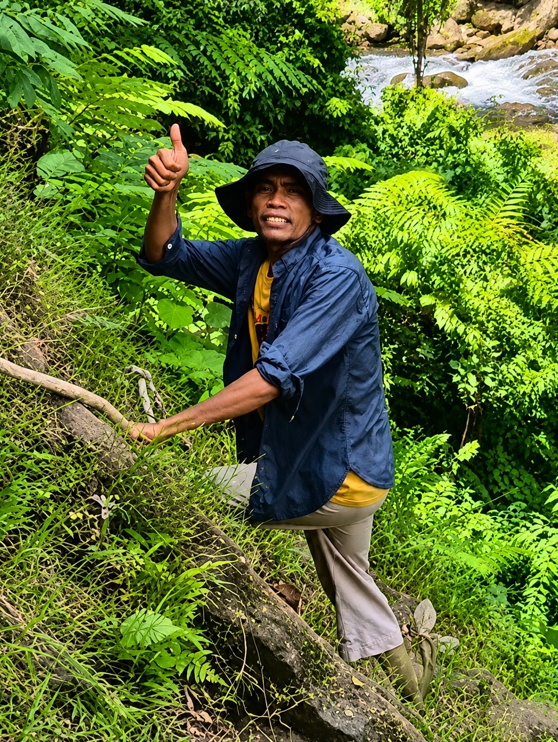 A medium shot captures a man with brown skin and short, dark hair wearing a dark bucket hat, a dark blue button-up shirt over a yellow t-shirt, and light brown pants. He is walking up a grassy incline next to a large, fallen tree trunk. He is looking directly at the camera with a wide smile and his right thumb is raised in a "thumbs up" gesture. The background is filled with lush green foliage and a waterfall with white, rushing water. The scene appears to be outdoors in a tropical environment.