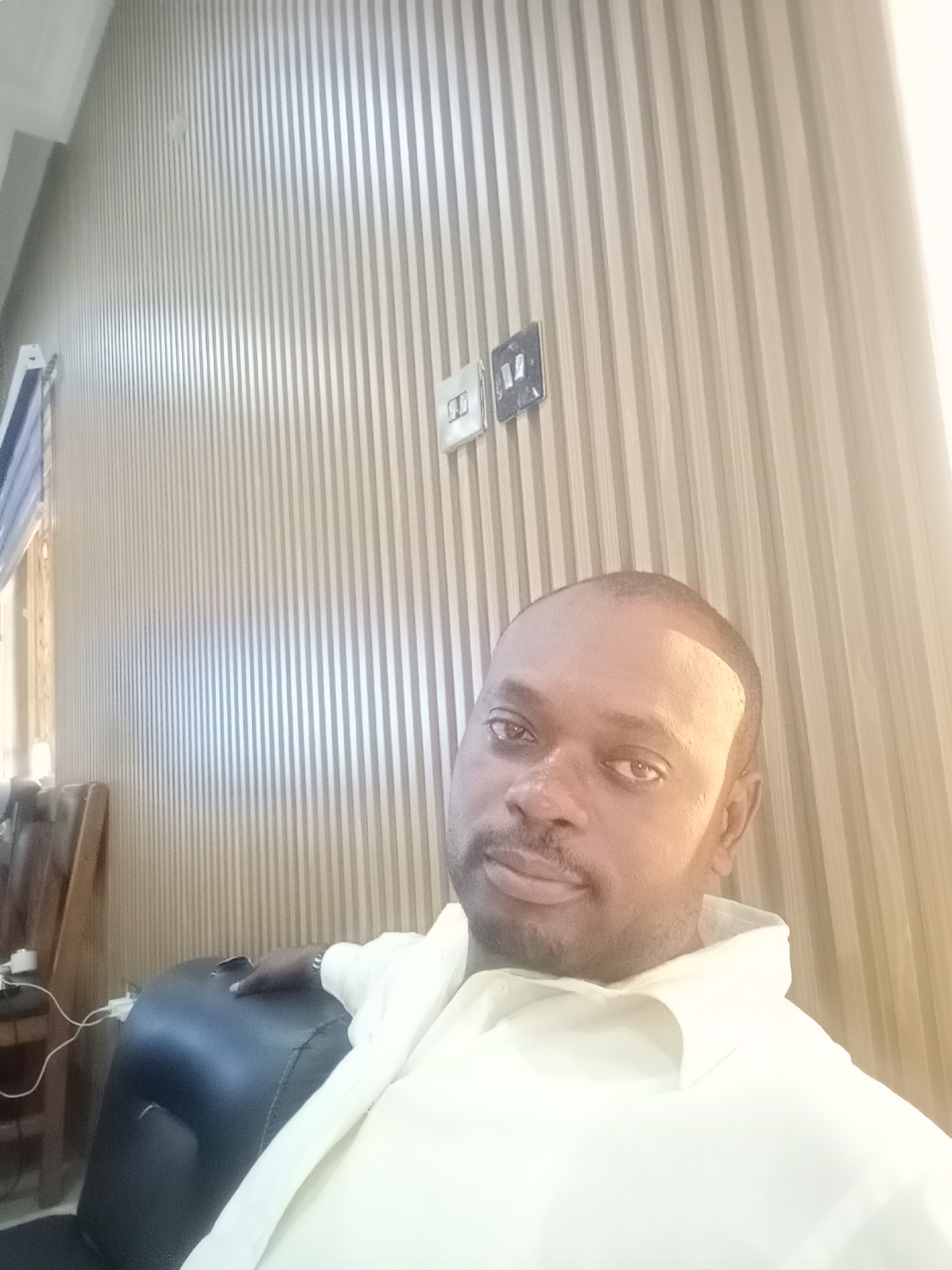 The image depicts a man with short dark hair and facial hair, wearing a white collared shirt. He appears to be seated in a black leather chair. His right hand rests on the chair's armrest. The background consists of a wall with vertical, alternating light and dark panels, and two light switches. Part of a window is visible on the left side, with an open blind. The location is speculated to be Gwarinpa, Nigeria, although this cannot be verified from the image alone.