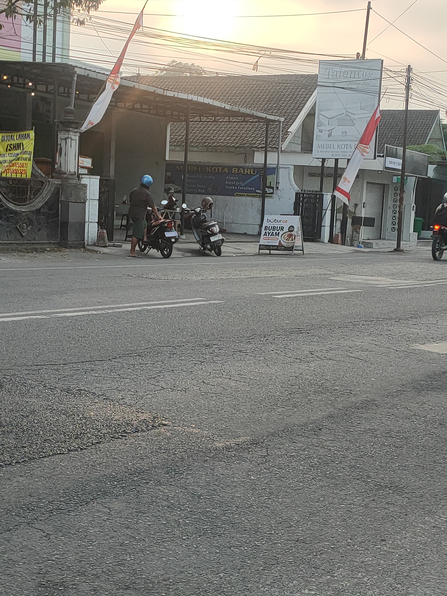 The image shows a street scene in Kanigoro, Indonesia, near a furniture store and a food stall. In the foreground, the asphalt road has faded white lane markings. On the left side of the street, there is a yellow sign advertising "Dijual Lahan" (Land for Sale). Behind the sign, a building with a covered outdoor area can be seen. In front of this building, two men are standing next to a motorcycle, and another motorcycle is parked to the right. A third motorcycle, with a rider, is visible further down the street on the right.
To the right of the first building, a larger establishment with a sign reading "Talent@" and "MEDEL KOTA BARU" is present. This building has a tiled roof and an Indonesian flag displayed prominently. Next to it, a white A-frame sign advertises "bubur.ae BUBUR AYAM" (Chicken Porridge). Further down the street, there appears to be another building with a covered walkway and a green trash bin. The sky above is overcast with the warm hues of either sunrise or sunset. The overall impression is of a typical street scene in a moderately busy area.