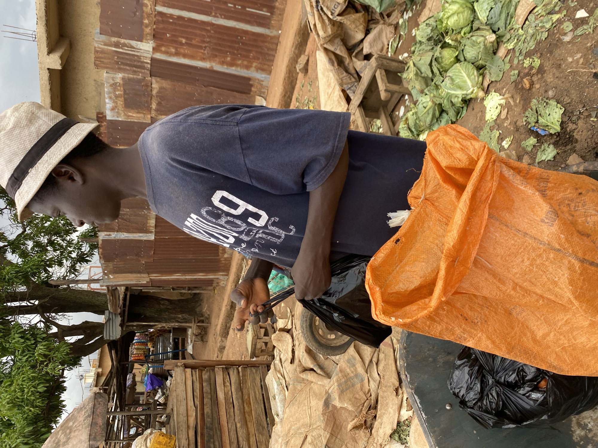A young person wearing a straw hat and a dark blue t-shirt is the main subject of this image, which appears to be taken in an outdoor market setting in Bukuru, Nigeria. The person is seen from the side, with their head bowed slightly as they hold the handles of a black plastic bag. This bag is partially contained within a larger, crinkled orange mesh bag, which is draped over a dark surface.
The background reveals a rustic environment with elements typical of a marketplace. There are structures made of corrugated metal and wood, suggesting stalls or makeshift shelters. Piles of green cabbage are visible on the ground, some appearing somewhat wilted or damaged. The ground itself is a mixture of dirt and what looks like discarded materials. A large tree with lush green foliage is present to the left of the frame, its branches extending into the upper portion of the image. The sky is overcast, indicating it might be a cloudy day.
The overall scene suggests a moment of activity related to collecting or sorting items, possibly waste or produce. The person's posture and focus on the bag imply a task being carried out. There are no clear expressions visible on the person's face, but the act of handling the bags suggests a purposeful action. A close-up crop of the person's t-shirt shows a white graphic with the letters "B" and other indecipherable characters, but no other discernible text is immediately apparent in the scene. The lighting is diffused, consistent with an overcast sky. The setting appears to be a somewhat unkempt area of the market, with debris and rough-hewn structures.