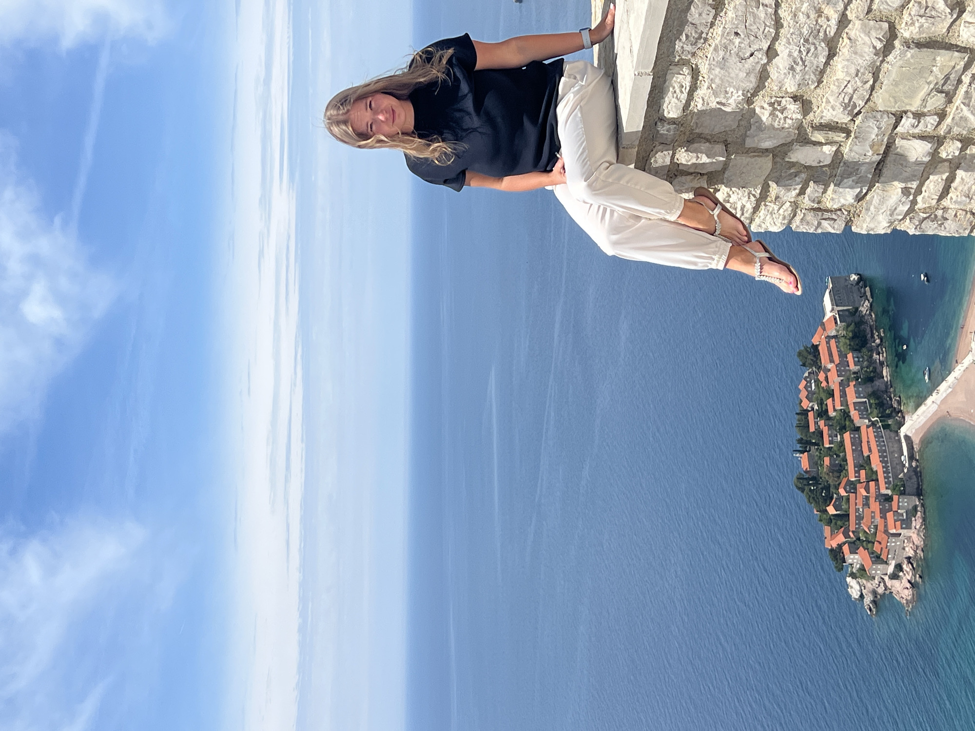 A woman sits on a stone wall, overlooking the sea and an island in Sveti Stefan, Montenegro. The woman has long blonde hair and wears a black t-shirt and white pants. She is smiling and looking toward the camera. Her legs are dangling over the wall, and she is wearing sandals. The stone wall is made of light-colored stones, and beyond it, the sea is deep blue. An island with red-tiled roofs is visible in the background to the right of the image. The sky above is blue with some clouds.
