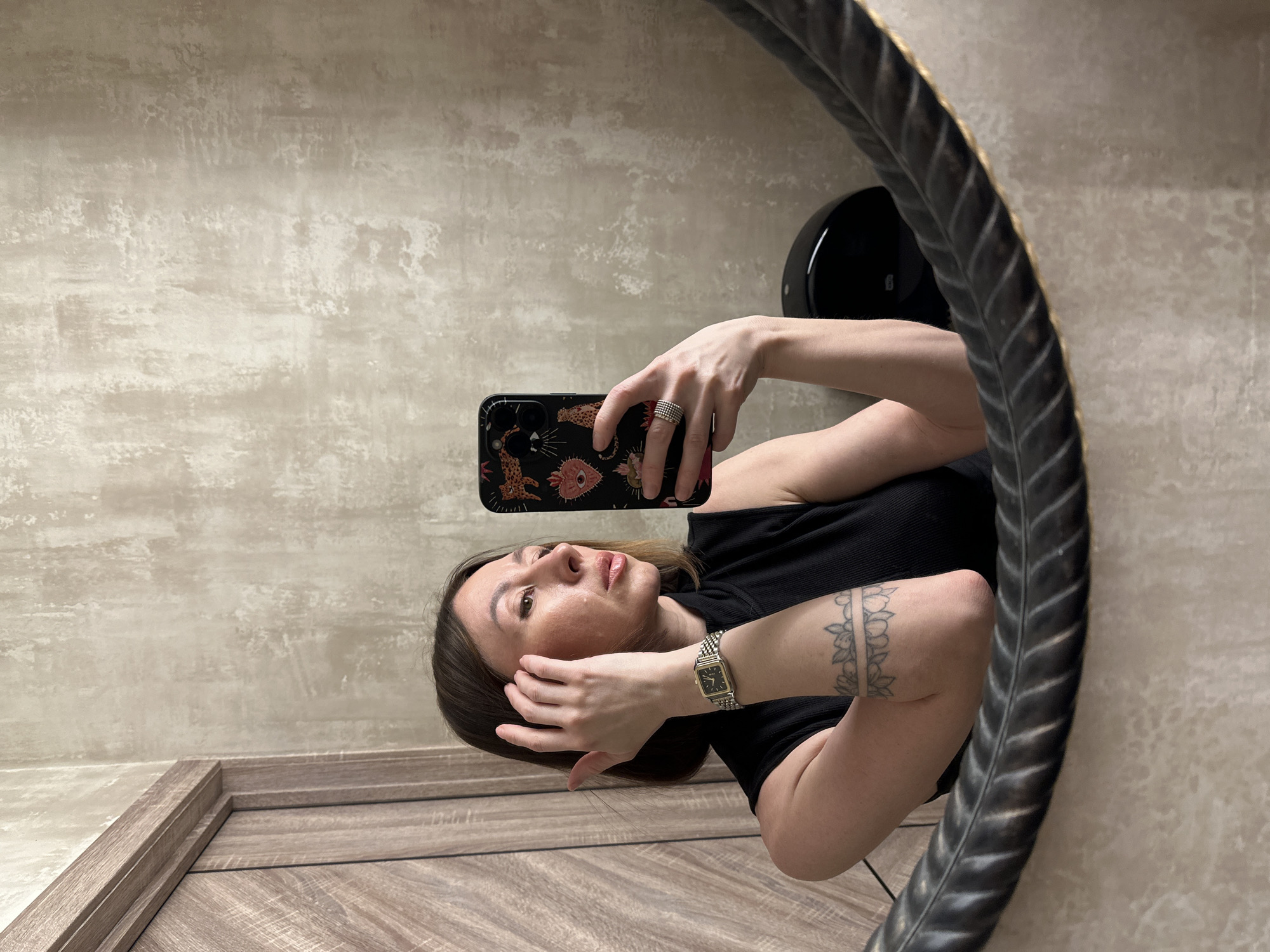 A woman is taking a selfie in a mirror. She is holding a smartphone with a patterned case in her right hand. She is wearing a black top and has tattoos on her left arm. She is wearing a watch on her left wrist and rings on her right hand. The mirror has a dark, textured frame. The background includes a beige wall and a wooden door or cabinet. A black object is partially visible on the wall behind her. The location is potentially in Izhevsk, Russia, although there is no definitive visual evidence to confirm this.