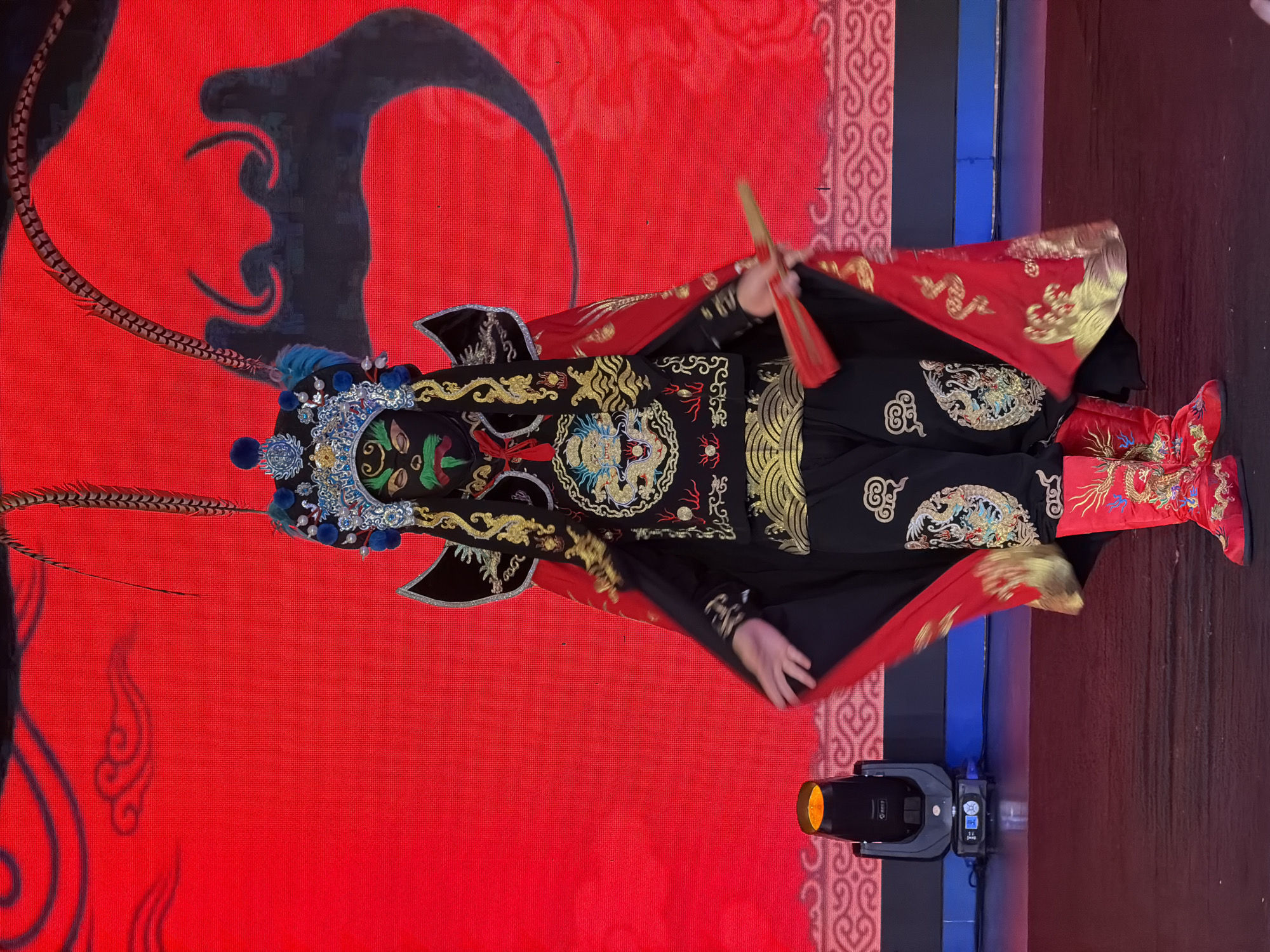 A performer in traditional Chinese opera attire, featuring a black costume with gold embroidery of dragons and clouds, a red cape, a detailed headdress with feathers and blue accents, and vibrant red boots, stands center stage. The performer's face is painted with a black base and green and red facial features, typical of a warrior character. They are holding a small red fan in their right hand and are positioned in front of a large red screen displaying intricate black and red decorative patterns resembling stylized clouds or mythical creatures. The stage floor is dark. To the left, a spotlight is visible. The setting appears to be a performance venue in Yuzhong District, China.