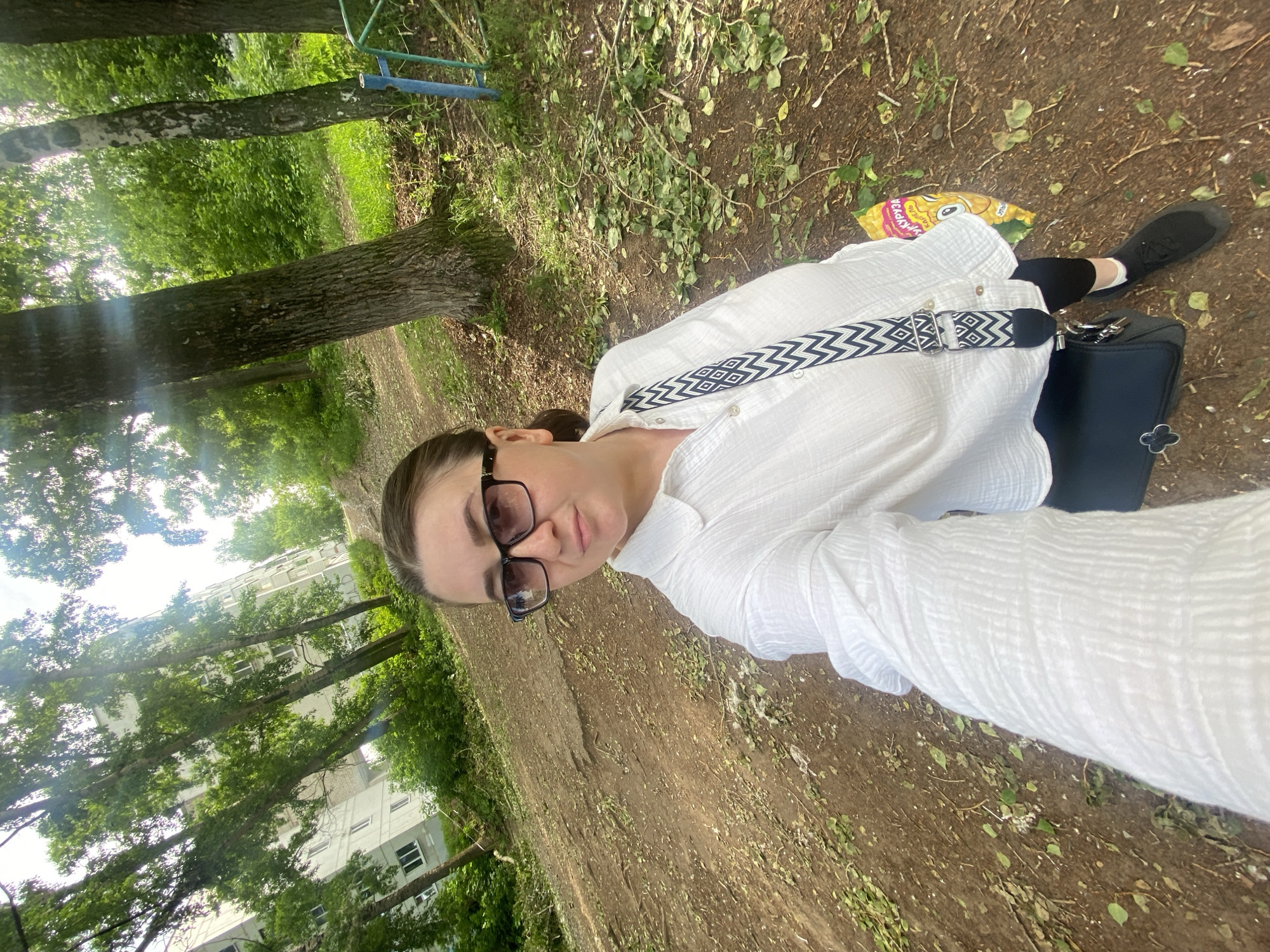 A medium shot, slightly rotated, captures a young woman taking a selfie outdoors in what appears to be a green area or park in Chaikovsky, Russia. The woman, positioned in the foreground and center-left, looks directly at the camera with a neutral expression. She wears clear-framed glasses, a loose-fitting white long-sleeved shirt with a textured fabric, and dark-colored pants. A black shoulder bag with a distinctive white and black chevron/geometric pattern strap hangs across her body. Her dark hair is pulled back.
The background features a natural, somewhat unkempt setting. The ground is a mix of dirt and patches of green grass, with scattered fallen leaves and twigs. Tall, mature trees with visible trunks and lush green foliage dominate the midground and background, suggesting a wooded or park-like environment. Sunlight creates bright spots and lens flare, indicating a daytime setting. Through the trees in the far background, a multi-story residential building with multiple windows is visible, suggesting an urban or suburban location. A blue metal structure, possibly part of playground equipment or a fence, is partially visible in the upper right background.
On the ground near the woman's right foot, a crumpled yellow bag of chips is visible. Cyrillic text is clearly printed on the bag, with the word "КУКУРУЗА" (KUKURUZA, meaning "corn") readable, identifying the snack. The woman's black shoes are also visible on the dirt ground. The overall scene suggests a casual moment captured in a public outdoor space during daylight.