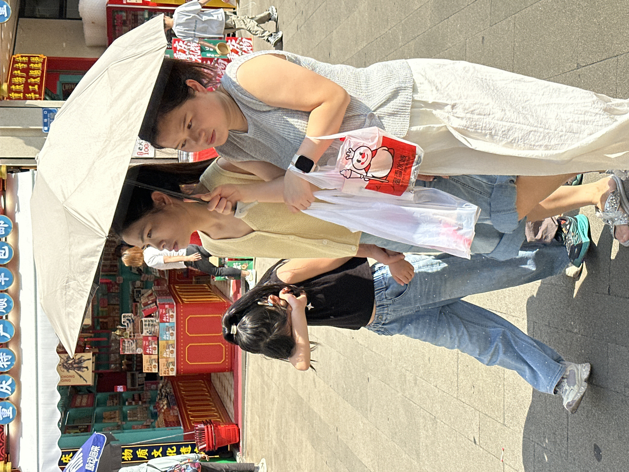 This media file captures a vibrant street scene, likely a commercial or tourist area, in bright daylight. The main subjects are three females walking together. In the foreground, an adult woman, wearing a grey knitted sleeveless top and a long white pleated skirt, holds a large, light-colored umbrella for shade. She carries a clear plastic bag containing a red item with a cartoon figure and text, and another white plastic bag. A black smartwatch is visible on her left wrist.
Beside her, another female, possibly an older teenager or young adult, dressed in a yellow long-sleeved cardigan over a light-colored top, also holds the umbrella handle. Ahead of them walks a younger girl, wearing a black crop top and light blue jeans speckled with white dots, her dark hair pulled back with a light-colored clip. She has her hand near her face. All three appear to be moving along a paved sidewalk, with strong shadows indicating the sunny weather.
The background reveals a bustling environment with various shops and market stalls. Colorful signs with Chinese characters are prominent. Visible text includes "蜜雪冰城" (Mixue Bingcheng), a popular ice cream and tea chain, on the plastic bags, confirming the brand. Other signs read "重庆特产传播" (Chongqing Specialty Products Promotion) and "手工火鍋底得" (Handmade Hotpot Base) along with "CHONG QING," consistent with the provided Yuzhong District, China, location context. Other fragmented text includes "脉动回来" (Maidong Huilai), "物质文化遗" (Material Cultural Heritage), "老火," and the number "100." The overall atmosphere is that of a casual outing or shopping trip on a warm, sunny day.