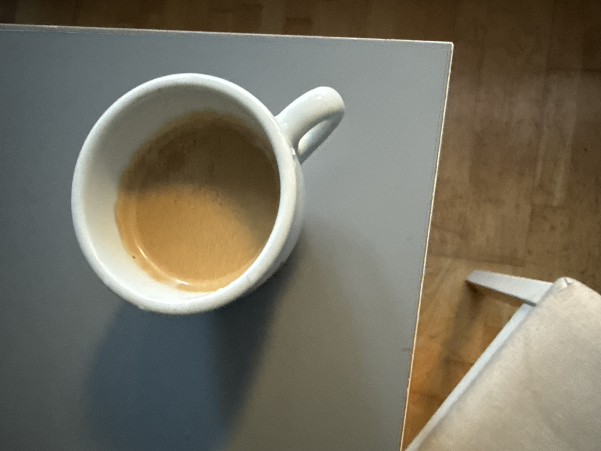 An overhead shot shows a white espresso cup filled with coffee, casting a shadow to its left on a grey table. The cup is positioned towards the upper right of the frame. The table surface is smooth and grey, with a slightly lighter grey edge. To the lower left of the image, a portion of a light-colored upholstered chair is visible. The background consists of a wooden floor with visible grain. The image does not provide any information about the city of Wrocław, Poland.