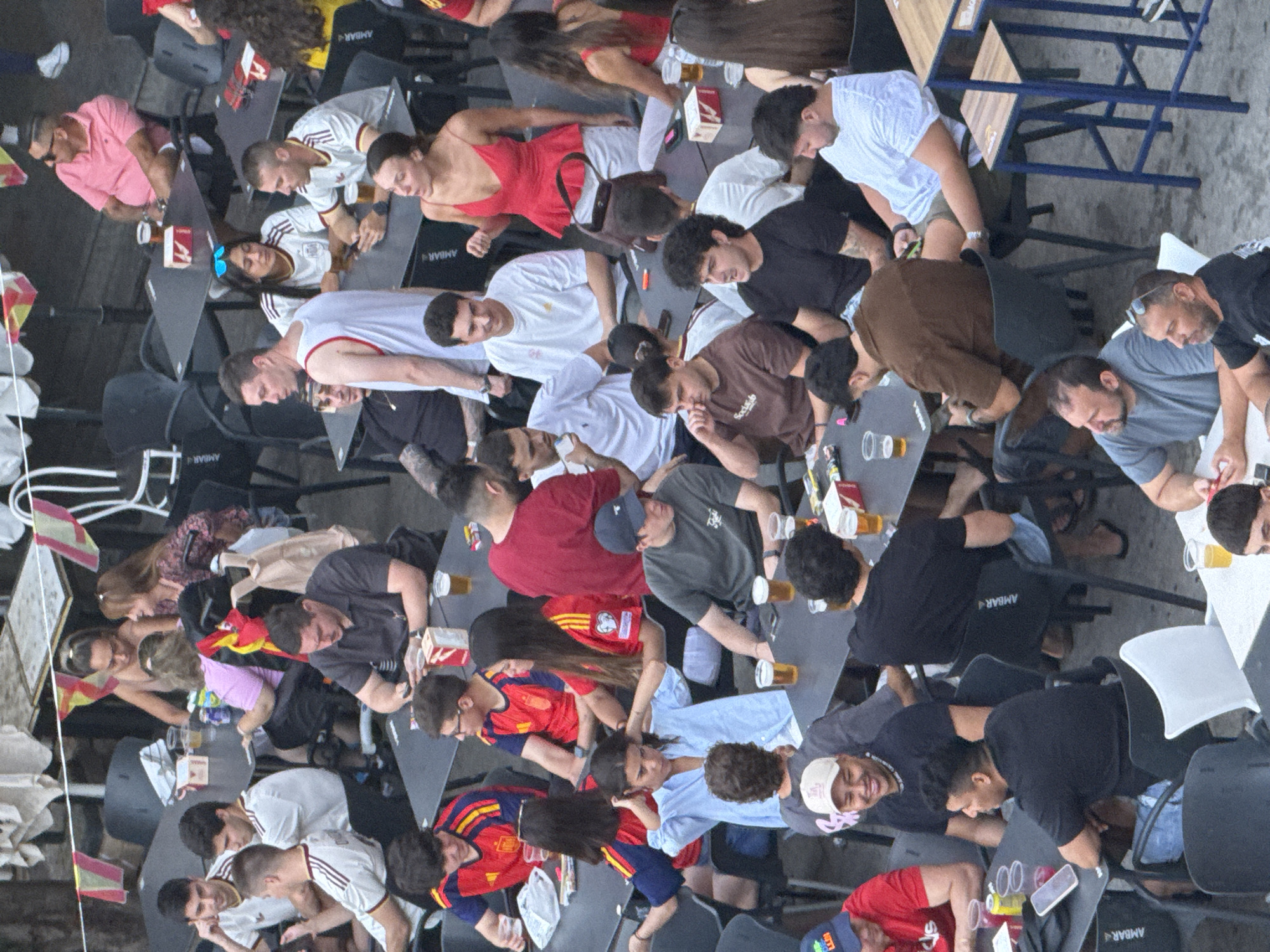 This high-angle, medium shot captures an outdoor seating area of a restaurant or bar in Las Rozas de Madrid, Spain. Approximately twenty individuals, appearing to be young adults and adults, are seated at various tables. Most are dressed casually, with some wearing Spain national football team jerseys. The atmosphere suggests a social gathering, likely related to a sporting event, given the jerseys and the presence of drinks on the tables. The environment is characterized by dark tables and chairs, typical of outdoor dining establishments. Red and yellow flags, possibly representing Spain, are visible strung above some of the tables, further reinforcing the context. The scene is populated by individuals engaged in conversation, observing their phones, and consuming beverages.