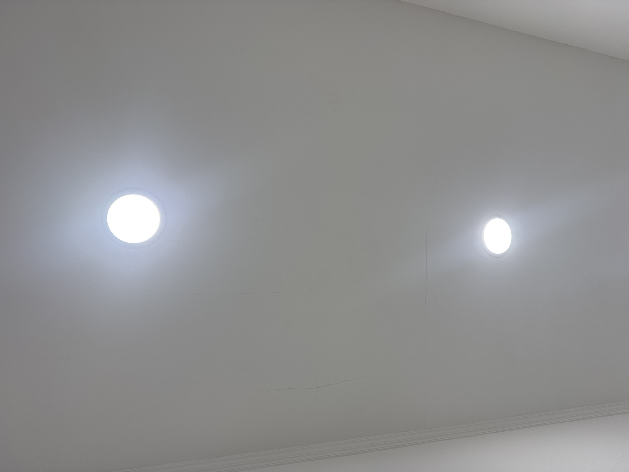 The image captures an interior ceiling, painted a light, neutral color, likely white or off-white. Two circular recessed lights, or downlights, are prominently featured, glowing brightly with white light and casting a soft illumination on the surrounding ceiling surface. One light is positioned towards the upper left, and the other is towards the upper right of the frame.
A decorative architectural molding, possibly a crown molding or coving, runs along the bottom edge of the image, indicating the juncture of the wall and ceiling.
Several hairline cracks are visible across the ceiling surface. A noticeable, thin horizontal crack extends across the lower central area of the ceiling, with a smaller vertical crack branching downwards from it. Other subtle, linear cracks are also visible in various parts of the ceiling, particularly towards the top edges and in the central region between the two lights. These suggest minor structural or cosmetic imperfections in the ceiling's finish.
The scene is static, with no people or activities taking place. The artificial lighting suggests an indoor setting, and no specific time of day is discernible from natural light. No visible text is present in the image. The depicted location is an interior space in Kuta Selatan, Indonesia.