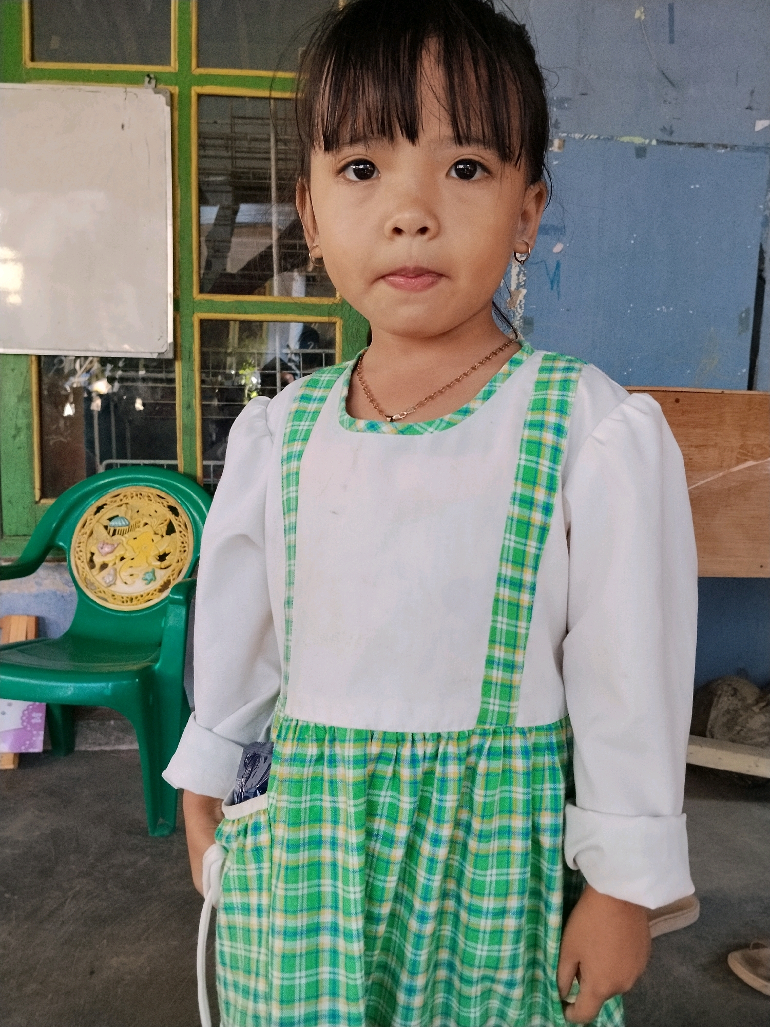 A young girl with dark hair and bangs stands in front of a blue wall and a green window frame. She is wearing a white long-sleeved dress with green and white plaid trim. The dress has a white bib and a plaid skirt. Her hands are in her pockets. To her left is a green plastic chair with a circular yellow design. A whiteboard is visible on the left side of the frame. The scene appears to be outdoors in a casual setting in Panggungan, Indonesia.