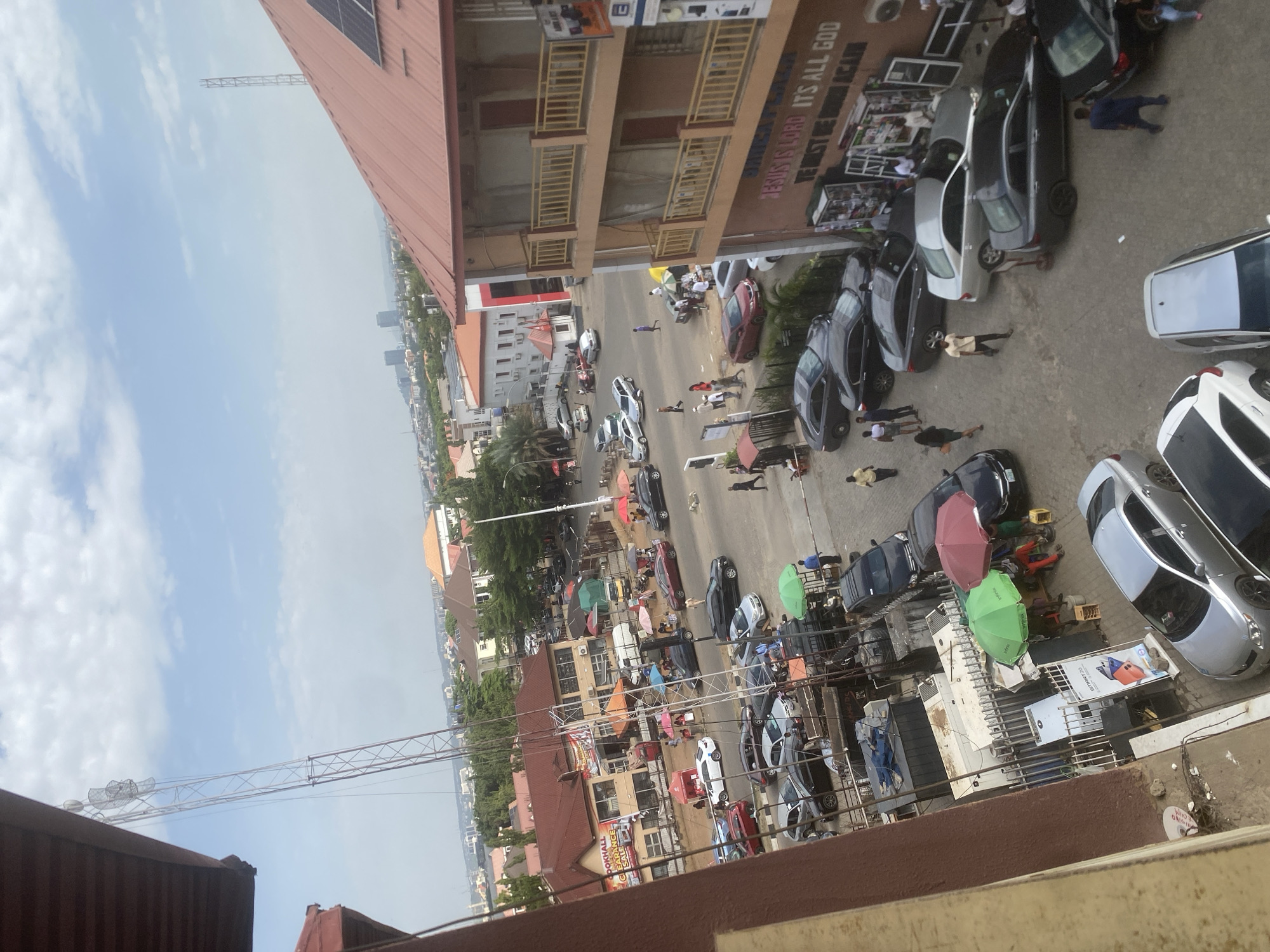 The image shows an elevated view of a street in Abuja, Nigeria. The street is lined with multi-story commercial buildings. Several cars are parked along the side of the road. Numerous pedestrians are present, some carrying umbrellas. Street vendors with stalls or tables are visible. Overall, the scene portrays a busy urban environment.