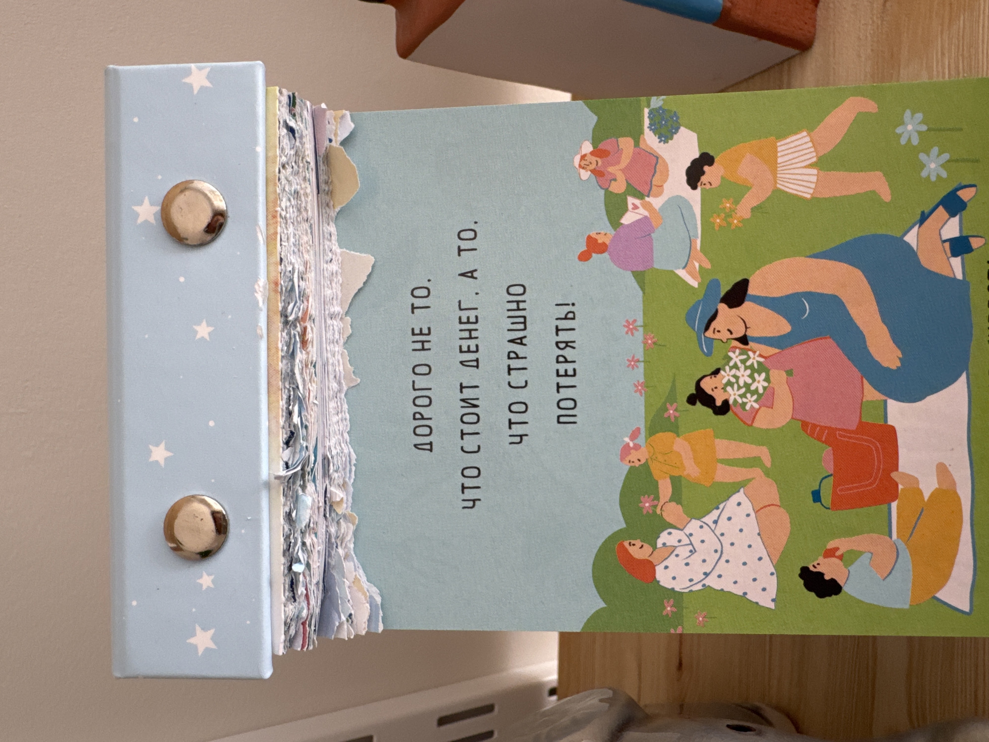 The scene is captured indoors in Izhevsk, Russia, displaying a desktop calendar resting on a light brown wooden surface. The calendar's top binding is a light blue cover adorned with white stars and small white dots, secured by two prominent, circular, gold-colored metallic studs. Below the cover, a thick stack of previously used pages is visible, characterized by their rough, torn edges and varied light colors, suggesting a daily or weekly tear-off format.
The currently displayed page features a light teal-blue upper section transitioning to a vibrant green lower section, depicting a grassy outdoor setting with stylized flowers. Central to the page is black Cyrillic text which translates to: "Expensive is not what costs money, but what you are afraid to lose!"
The lower portion of the page illustrates a group of approximately eight stylized women and a young person, all with a flat, cartoon-like aesthetic, engaged in a picnic or gathering on a green lawn.
- In the upper left, a woman with red hair in a white polka-dot dress is seated, holding hands with a standing young girl in a yellow dress.
- In the upper right, two women are seated on a white blanket: one in purple attire looking towards another with a wide-brimmed white hat and pink dress. Small blue flowers are near their blanket.
- Towards the middle-left, a woman in a pink dress is holding a bouquet of white flowers.
- In the bottom left, a woman in a light blue top and yellow pants sits on a white blanket, eating a red apple, with a red picnic basket and a blue bottle beside her.
- In the bottom right, a woman in a blue dress and a wide-brimmed blue hat is seated on a white blanket, holding white flowers, and wearing blue sandals.
- A young person in a yellow t-shirt and white striped shorts is walking towards the group from the lower right, carrying orange flowers. The green lawn is dotted with small pink, orange, and blue flowers.
To the immediate left of the calendar, a portion of a white vertical object with dark vertical slats is partially visible. To the immediate right, a section of a brown object with a blue top, possibly a decorative item or furniture, is visible.