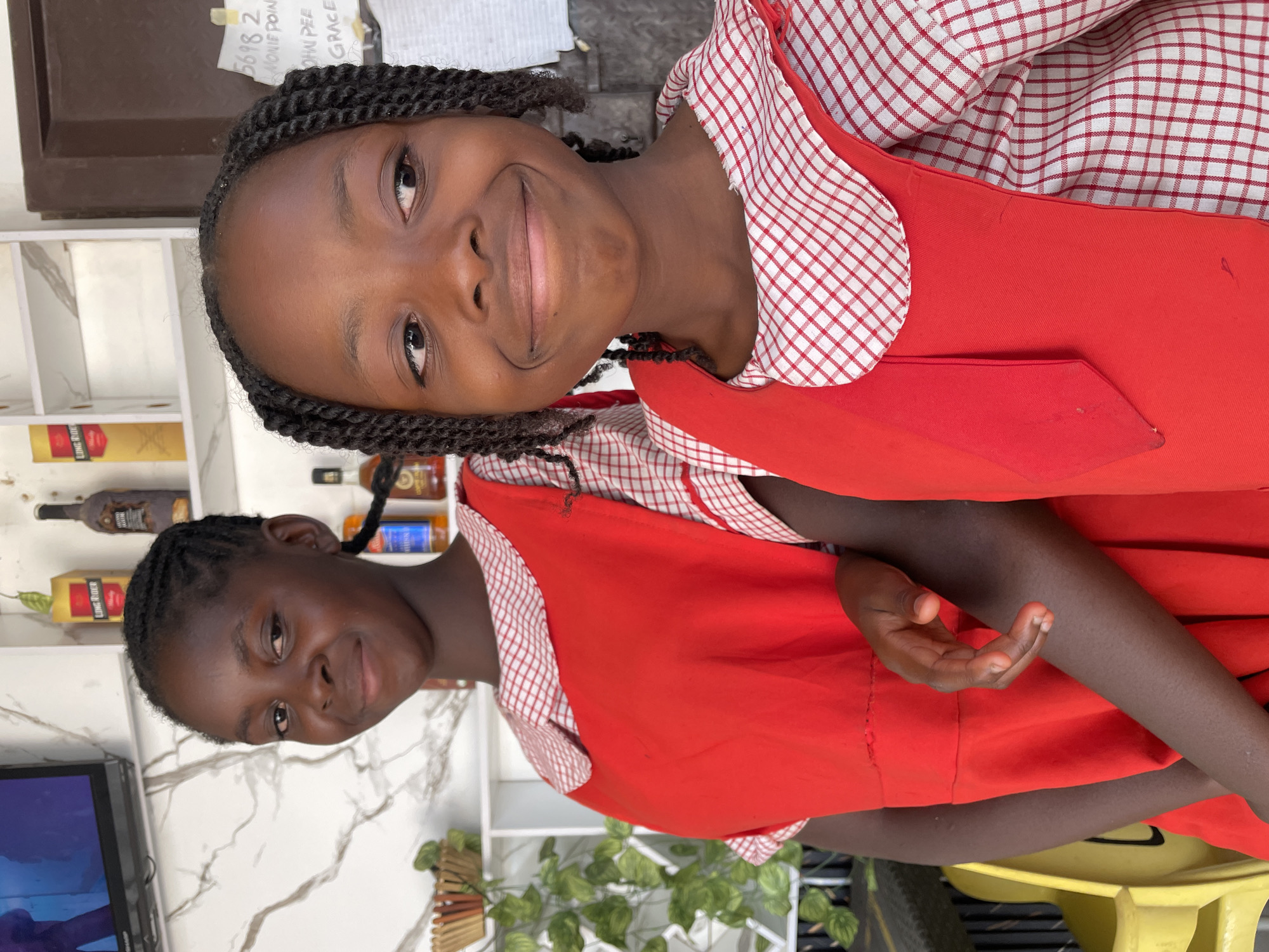 This image captures two young Black girls posing for a photograph indoors in what appears to be Barakin Akawo, Nigeria. Both girls are dressed in matching red dresses with white and red checkered patterns on their collars and sleeves, suggesting school uniforms. Their hair is styled in neat braids.
The girl positioned more prominently in the foreground has a wide, cheerful smile and looks directly at the camera. The second girl, standing slightly behind and to the right, also smiles gently. Both appear happy and relaxed.
The background reveals a white shelving unit stocked with various items, including several bottles. Labels partially visible on these bottles include "JACK DANIEL'S," "HENNESSY," and a clearer label for "LONG RIDER." A small black television screen is visible on the lower left, and some green foliage, possibly artificial plants, adorns a lower shelf. Part of a yellow plastic chair is seen at the bottom.
Taped to the wall or shelf above the girls' heads is a piece of paper with handwritten text, including "5698 2," "MONILE POINT," "OHN PEE," and "GRACE." The indoor lighting suggests the photo was taken during the daytime. The setting could be a home or a small local establishment.