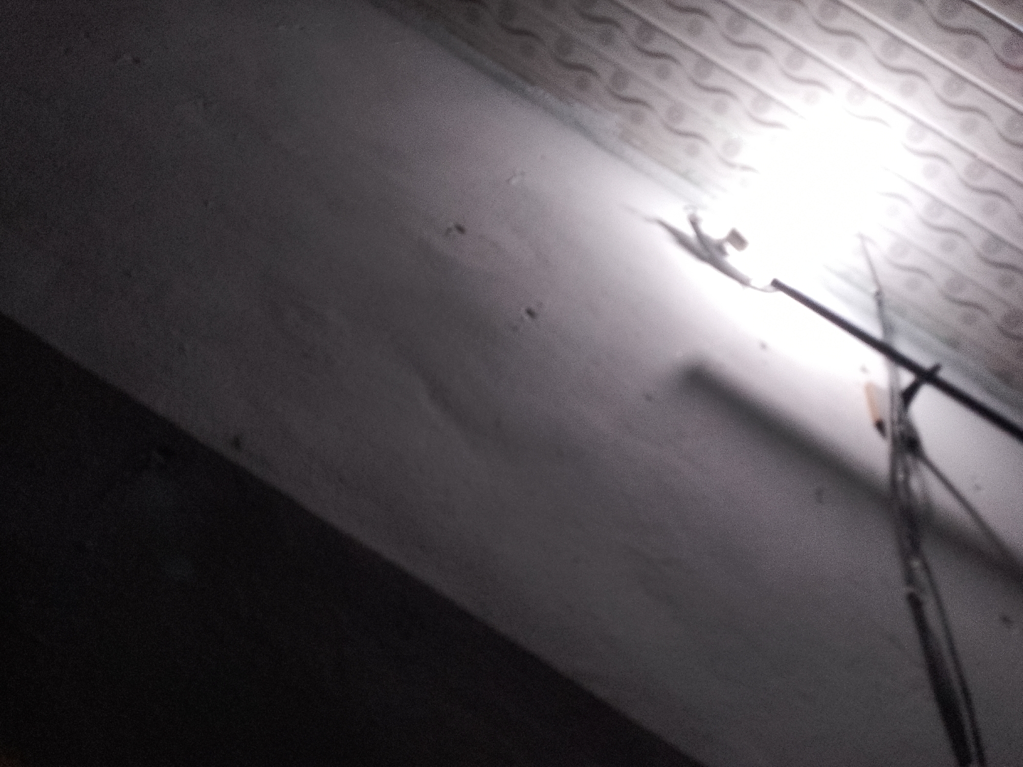 The image shows an interior view, likely a ceiling, with a light fixture and wires. The ceiling appears to be made of white, textured panels, possibly suspended ceiling tiles. A bright light source is visible, creating a strong glare. Black wires or cables are attached to the fixture and run downwards. The rest of the visible area is a grey, textured surface, possibly a wall or another part of the ceiling, with some faint shadows and dark spots. The overall impression is of a dimly lit, functional space. The image does not provide any contextual information that would indicate its location within Jauro Yinu, Nigeria.