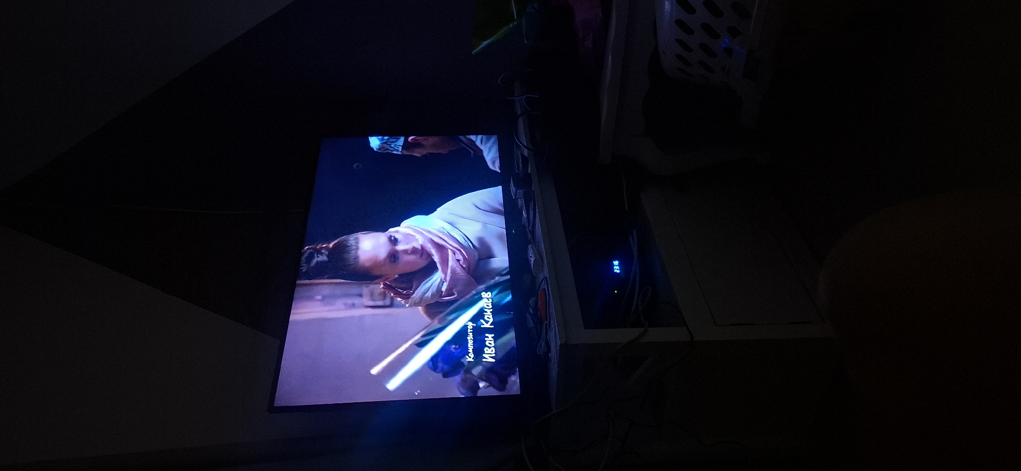 An indoor, dimly lit scene shows a television screen displaying a close-up of a woman with her hair pulled back in a bun, wearing a light-colored coat and a pink scarf. Text on the screen reads "Композитор" and "Иван Канаев" in Russian. The television is placed on a white stand, below which is a black electronic device displaying a blue digital clock reading "23:16". The background wall has a geometric design in dark colors. The scene does not provide any information or context related to the city of Yerocham, Israel.
