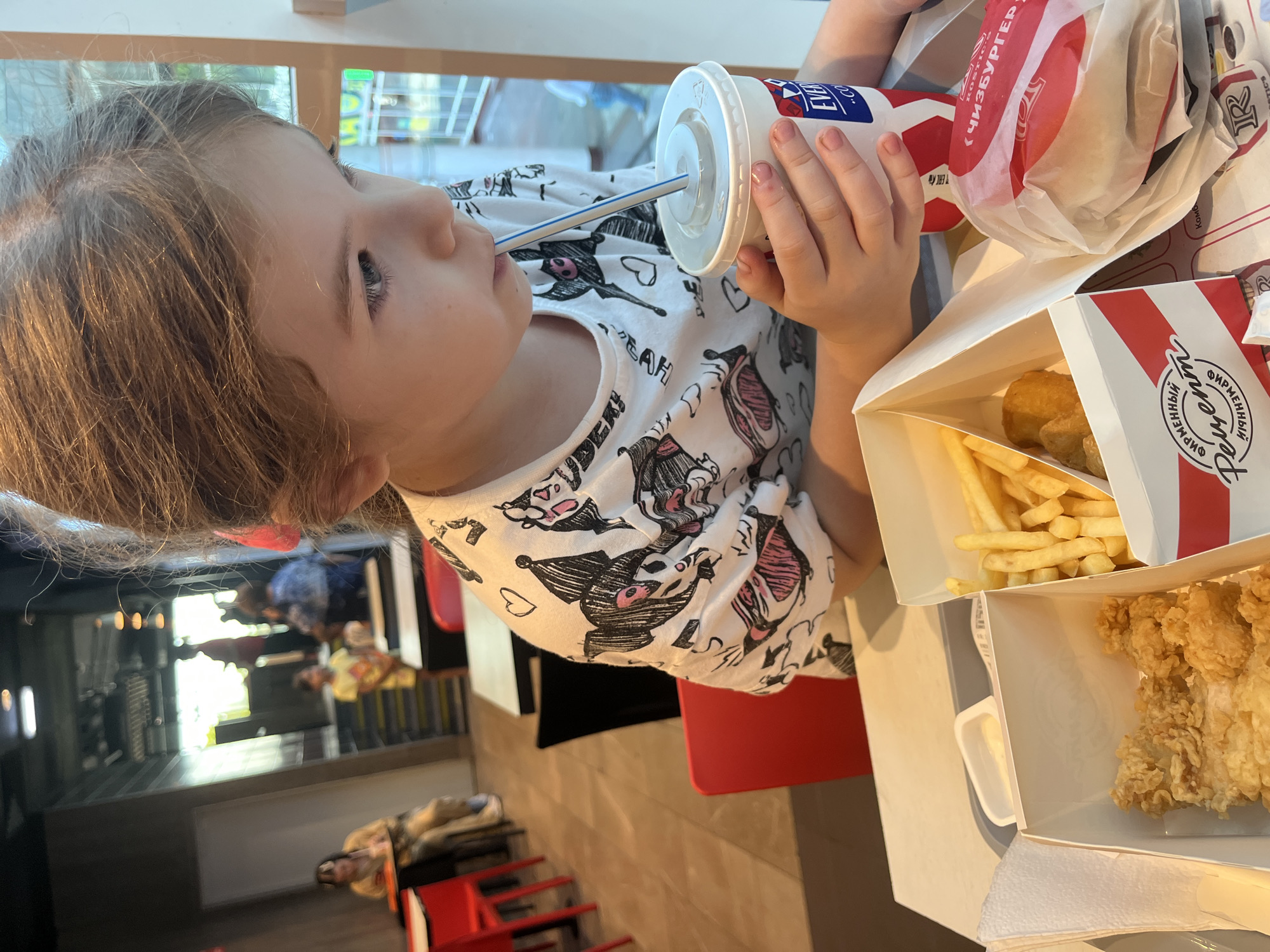 The image shows a young girl with blonde hair wearing a white t-shirt with a black and pink cartoon character print. She is sitting at a table, holding a white cup with a red and blue logo and a straw, from which she is drinking. The cup appears to be a beverage container from a fast-food establishment.
In front of her are several fast-food items, including a white paper bag with a red and white striped pattern, likely containing food. A white box contains french fries and pieces of fried chicken. Another white box is partially visible, also containing fried chicken. A small white container of dipping sauce is also visible.
The background suggests an indoor setting, possibly a fast-food restaurant. The girl's focus is on her drink, and her expression is neutral. The lighting indicates it could be daytime.
Visible text on the cup includes "EV," and on the bag and box, there is text in Cyrillic script, including "ЧИЗБУРГЕР" which translates to "Cheeseburger" and "ФИРМЕННЫЙ." The text and the food items are consistent with a fast-food meal. The scene appears to be in Sochi, Russia, as indicated by the location context.