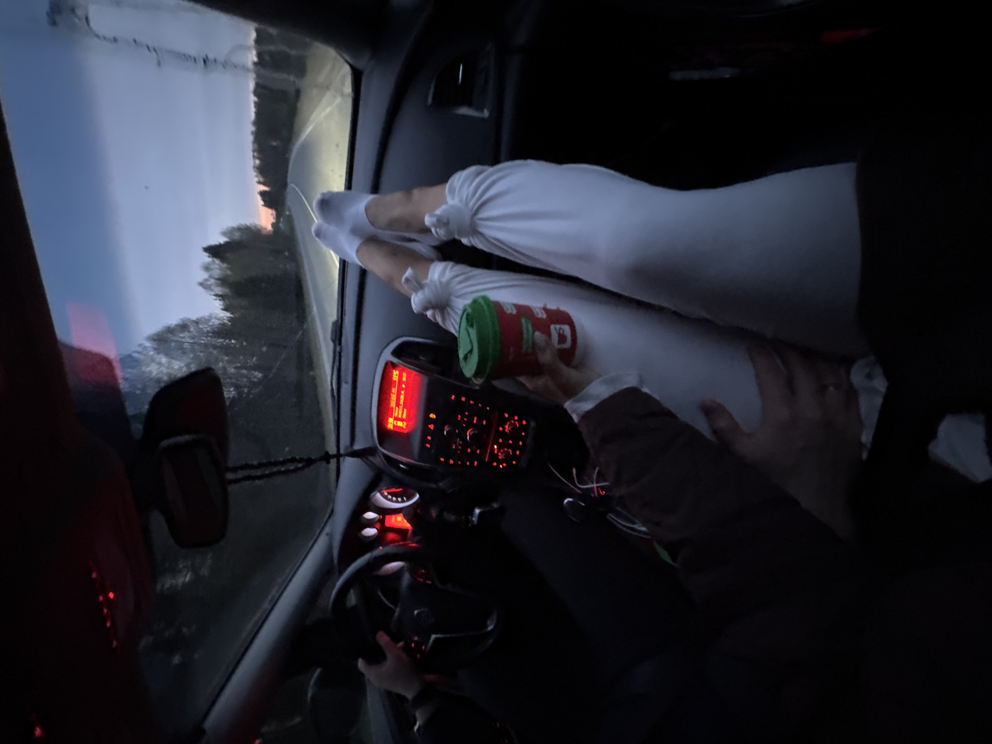 The interior of a car is shown from a slightly tilted and elevated perspective, likely taken by someone in the back seat. The dashboard and steering wheel are visible, with illuminated controls and a red display screen. A person in a dark jacket is visible holding a red and green container. A passenger has placed their legs, clad in white socks and pants, on the dashboard. The background shows a road leading into a forested area at dusk or dawn. It is unlikely to determine the precise location within the city of Полозово from the provided image.