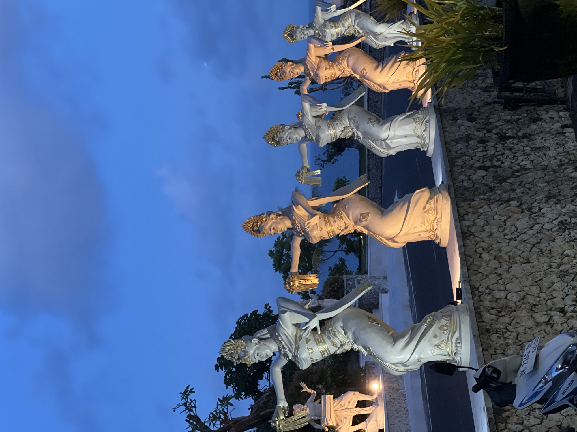 Captured at night in Kuta Selatan, Indonesia, the image features a series of Balinese dancer statues lined up alongside a road. The statues are light-colored with gold detailing and are illuminated by spotlights. They are set against a backdrop of trees and a body of water, likely the ocean. A parked white and blue Honda scooter is visible on the right side of the frame, with its license plate partially obscured. The sky is a deep blue color with a few clouds. The statues appear to be part of a decorative display along the roadside.