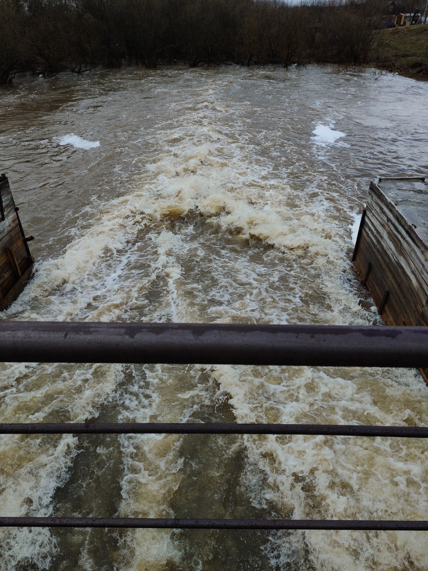 A high-angle shot depicts a turbulent, muddy river coursing through a concrete channel. The perspective is from a bridge, indicated by metal railings visible in the foreground, suggesting the location is within an urban area. The water churns with white foam, hinting at a strong current or recent heavy rainfall. On either side of the channel, there are wooden or concrete structures possibly functioning as retaining walls. The background reveals a thicket of bare trees and vegetation along the riverbanks, with a small building visible in the distance to the upper right. The overall tone is somber, due to the overcast sky and the color of the water. The scene is in Unknown, Unknown.