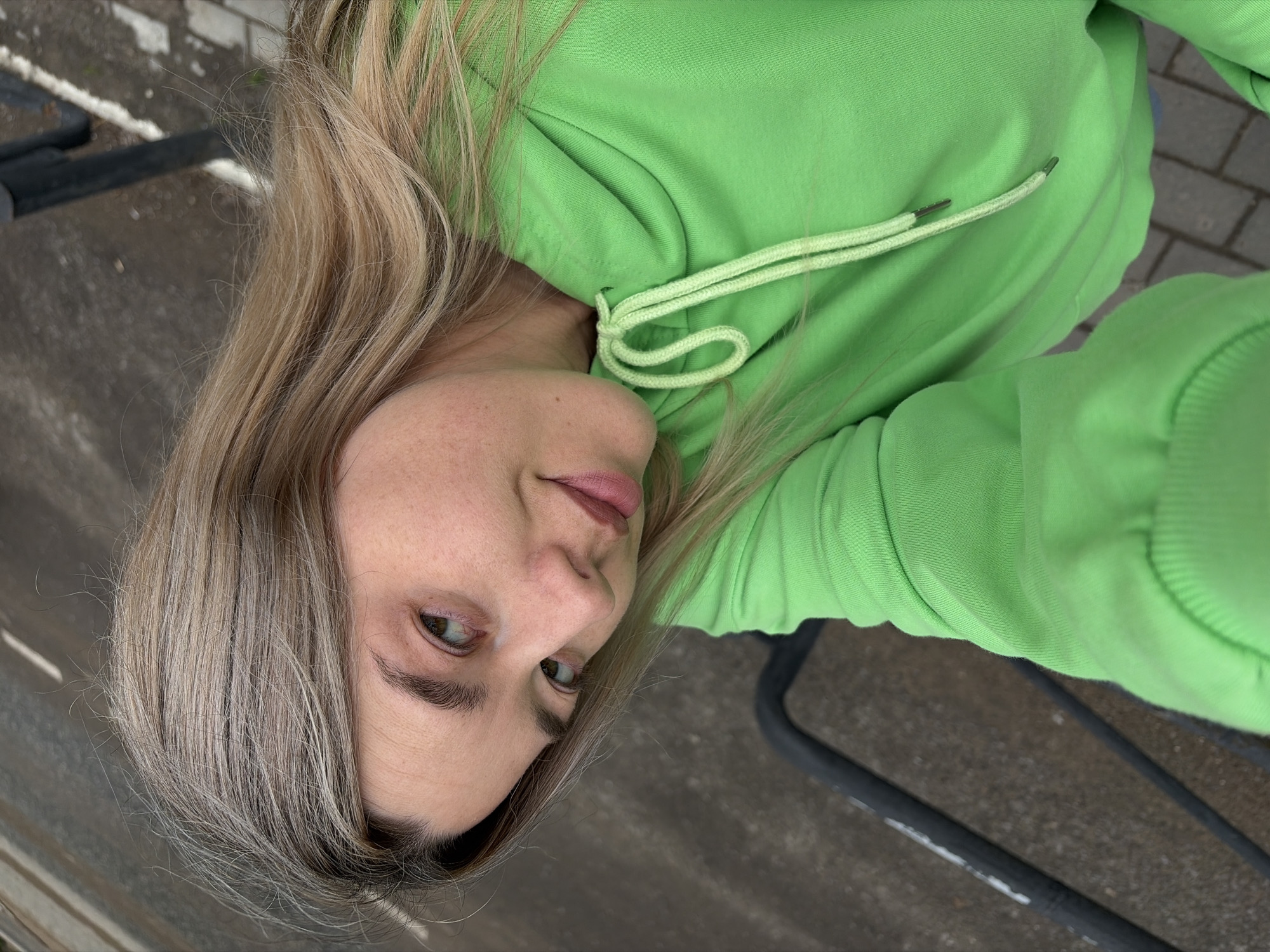 A tilted, close-up outdoor portrait features a woman with long, light blonde to ash-blonde hair, styled to fall over her shoulders. She is wearing a vibrant lime green hooded sweatshirt with matching drawstrings, which have silver-toned aglets. The woman looks towards the camera with a subtle, neutral expression and a slight smirk. Her eyes are light brown, and her lips have a soft pink tint. The image is captured from an upward angle, emphasizing her face and upper body.
The background, largely out of focus due to the shallow depth of field, shows a paved ground surface, possibly concrete or light-colored bricks, along with dark metal structures that resemble parts of outdoor furniture or railings. The natural lighting suggests it is daytime, possibly under an overcast sky or in a shaded area. This casual shot, taken in Chaikovsky, Russia, conveys a relaxed mood. There is no visible text in the image.