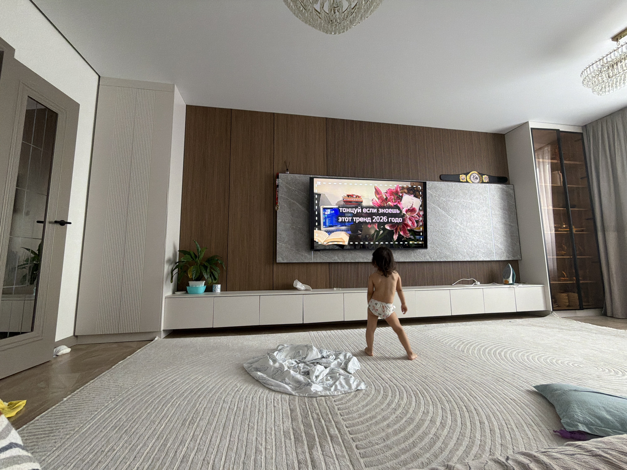 In a spacious, modern living room, a young child with dark hair and wearing patterned briefs stands barefoot on a large, plush, light-colored rug with a textured, concentric pattern. The child is facing away from the camera, towards a large flat-screen television mounted on a wood-paneled wall. The television displays a video with text overlayed in Russian, featuring images of flowers and what appears to be a book. To the left of the television is a gray, marble-patterned console. Below the television, a long, white modular cabinet unit runs along the wall, with several closed doors. On top of this unit, to the left of the television, a potted plant sits in a blue container, beside a smaller plant and a white object. Further to the left, a tall, light-gray cabinet with vertical ribbed detailing stands next to a door with glass panes and a black handle. To the right of the television, a decorative belt with a gold buckle is displayed on a ledge. In the background, a tall glass-doored cabinet filled with items can be seen, along with gray curtains. A crumpled light-colored blanket lies on the rug in front of the child, and a blue pillow is visible in the lower right corner of the frame. The room is illuminated by ambient light, with a chandelier visible in the upper part of the image. The overall impression is that of a contemporary, well-furnished residential interior, likely in a city like Astana, Kazakhstan, given the provided context.