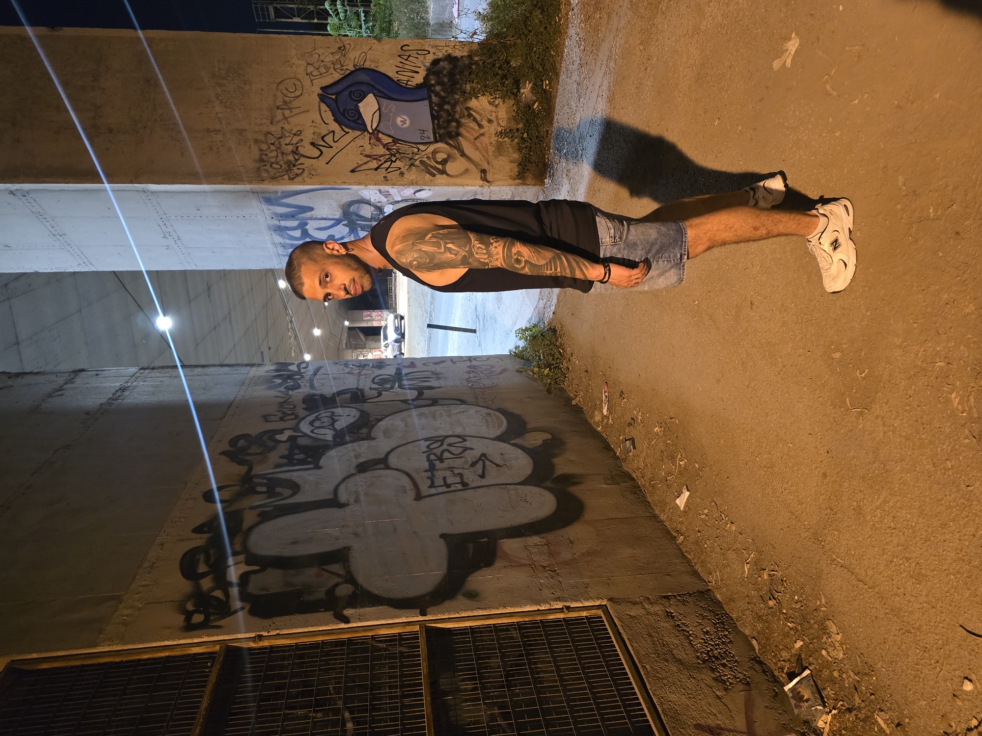 The image displays a male subject standing in an urban underpass or tunnel at night, identified as Athina, Greece, due to the visible Greek word "ΕΓΕΡΣ" (Egersi) incorporated into graffiti on the left wall.
The male, appearing to be in his late 20s or early 30s, stands in a three-quarter profile, looking towards the viewer over his left shoulder. He has short, closely cropped hair, a beard, and wears earrings in both ears. His attire consists of a black sleeveless tank top, light blue denim shorts, and white New Balance sneakers. A full sleeve tattoo, detailing multiple figures and potentially a crown, covers his left arm. A dark wristband is present on his left wrist.
The setting is characterized by concrete walls and large concrete pillars supporting an overhead structure, consistent with an underpass. Both walls are extensively covered with layers of graffiti. On the left wall, a prominent bubble-style graffiti includes the Greek word "ΕΓΕΡΣ". The right wall features a distinct blue, bird-like character with white facial elements, marked with the number "24" and partial text "ANIAN" or "ANIAS". A dark metal grate or fence structure is visible on the far left. The ground is unpaved, composed of dirt and gravel, with scattered litter including paper and plastic fragments. Sparse green vegetation grows along the base of the right wall. Multiple artificial light sources are visible in the distance under the overpass, generating distinct horizontal light streaks across the image. A white vehicle is discernible further into the passage. The man casts a prominent shadow to his right on the ground.