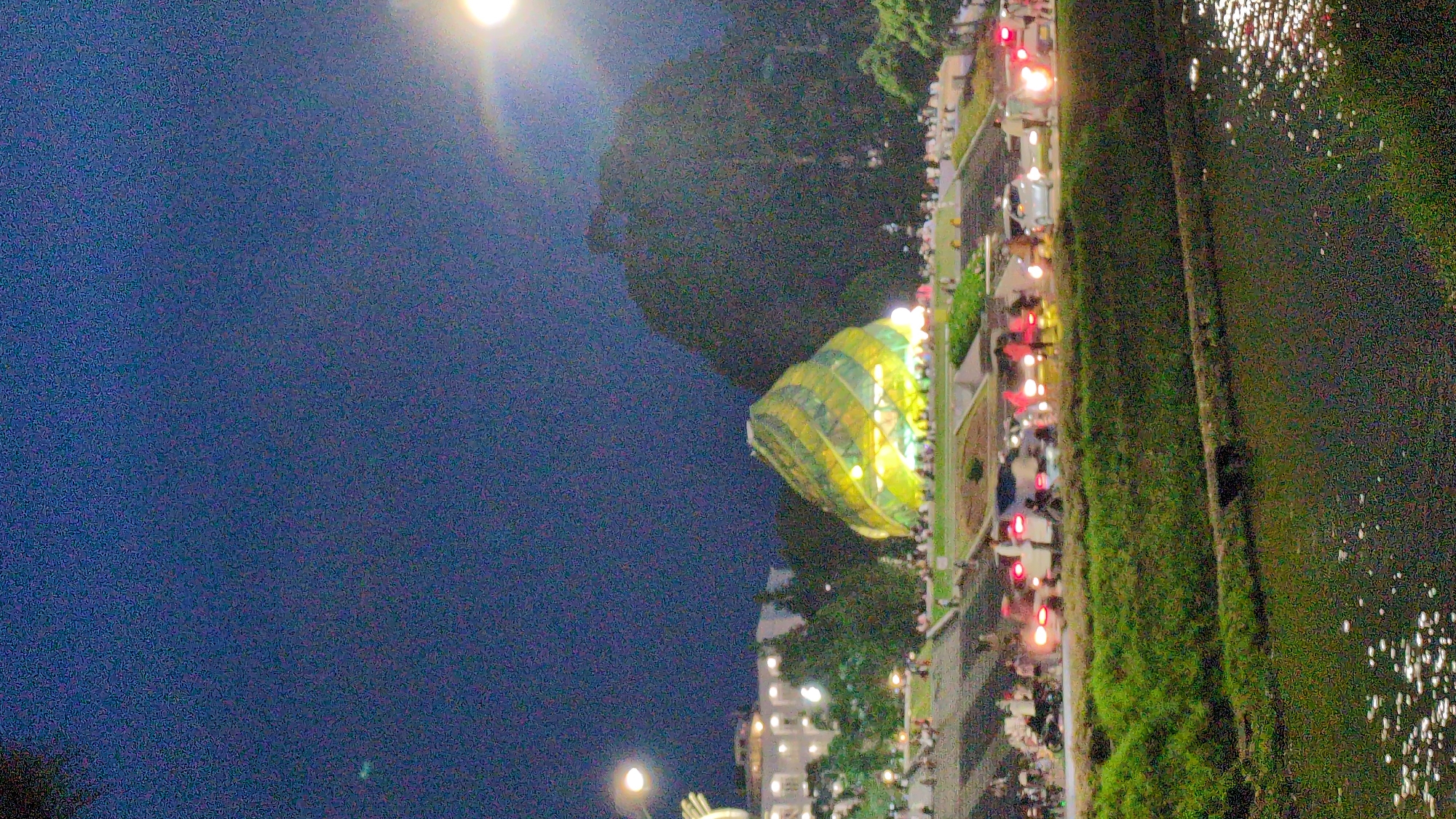 The image captures a nighttime scene in Da Lat, Vietnam, focusing on a unique, conical-shaped building illuminated with yellow and green lights. The structure appears to be a landmark, possibly a pavilion or exhibition center, situated in a park-like area with tiered landscaping and scattered trees. In the foreground, a body of water reflects the artificial lights.
A street runs parallel to the park, populated by a variety of vehicles including cars, motorcycles, and possibly scooters, all illuminated by their headlights and taillights. A crowd of people is visible on the street and in the park area surrounding the illuminated building, suggesting a public gathering or event. The background features a dark, tree-covered hill, characteristic of Da Lat's topography, and a bright light source, likely a streetlamp or floodlight, is prominent in the upper right corner. The overall atmosphere is vibrant and bustling, despite the darkness.