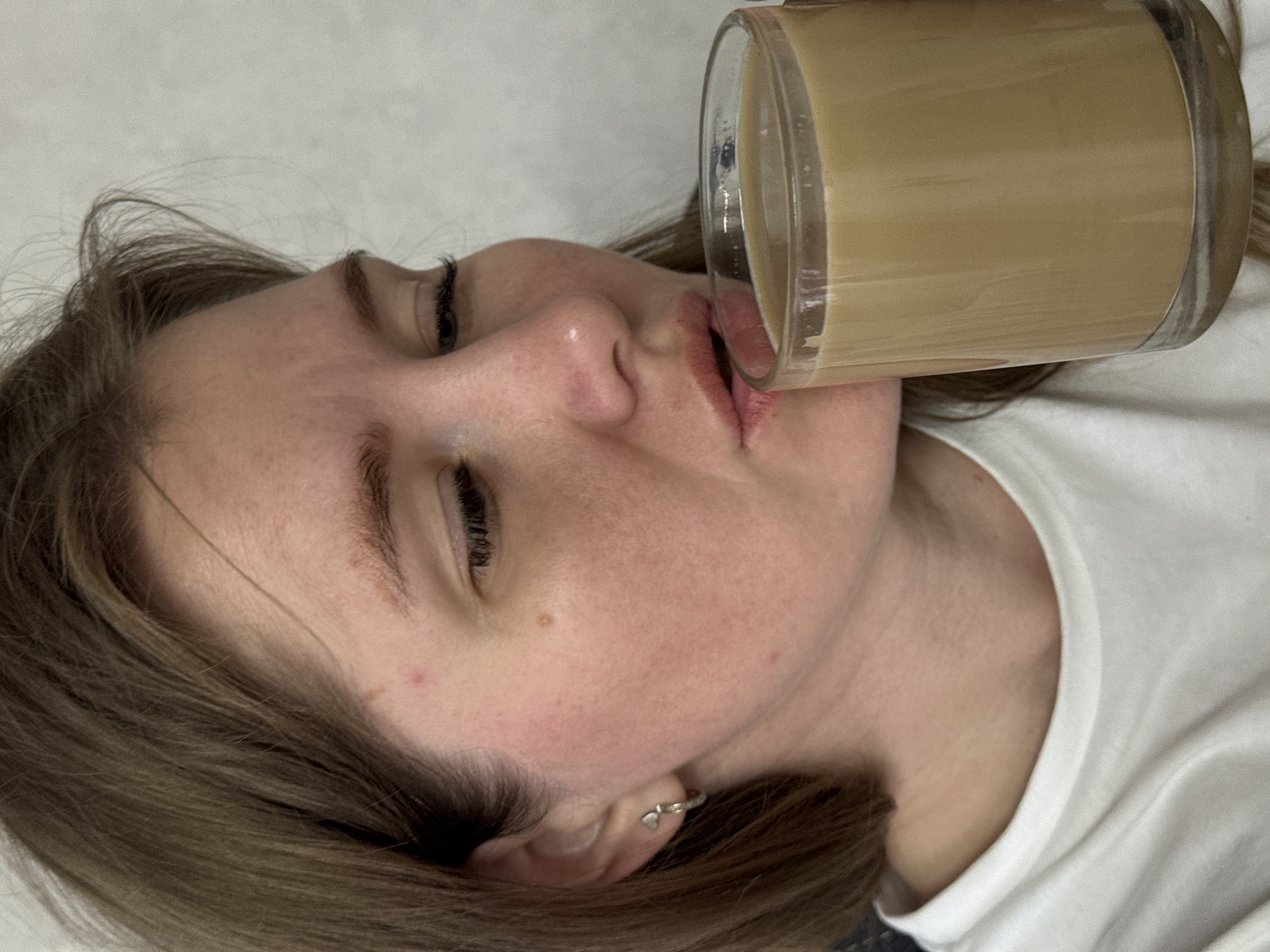 A woman is drinking from a glass. The glass contains a beige-colored liquid. The woman has brown hair, fair skin, and is wearing a white t-shirt. She has a small earring in her left ear. The background is a light gray or white surface. The image appears to be taken indoors. There is no information available to verify the location within the city of Gornyi (Dvurechenskoe S/P), Russia.