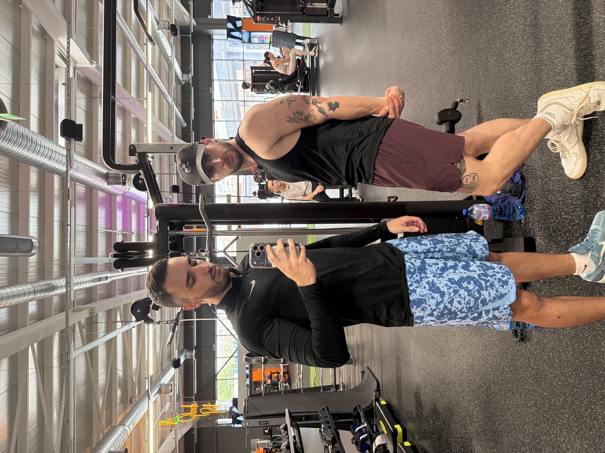 The image shows the interior of a gym. Two men are visible. One man, wearing a black tank top, maroon shorts, a grey baseball cap, and white shoes, is stretching his arms. He has tattoos on his arms and legs. The other man, wearing a black long-sleeve shirt, blue patterned shorts, and blue shoes, is taking a photo of the first man with his smartphone. Several exercise machines are visible in the background. There is a water bottle on the floor near the man in the maroon shorts. The location is likely in Plovdiv, Bulgaria, based on the user prompt.